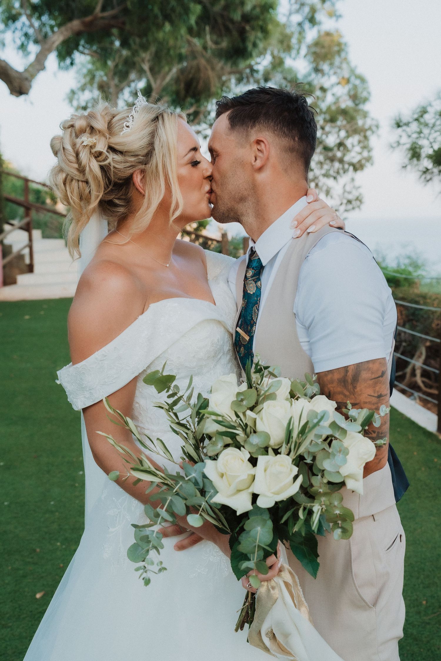 Romantic newlyweds share a kiss while holding a white and green bouquet during their outdoor garden wedding.