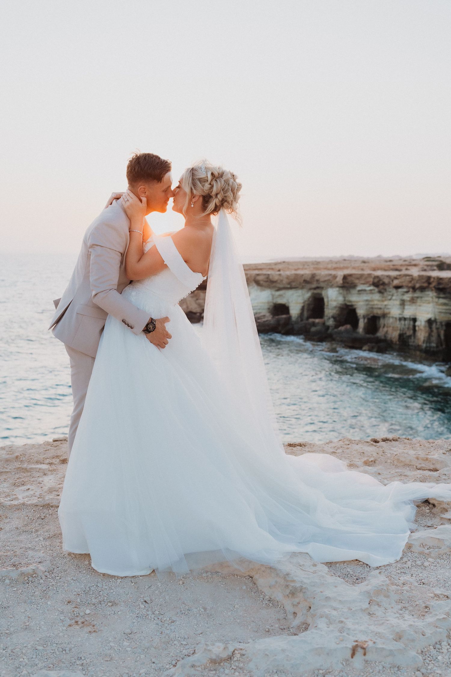 Newlyweds embrace on scenic coastal cliffs at sunset as bride's white veil and dress flow in the breeze.