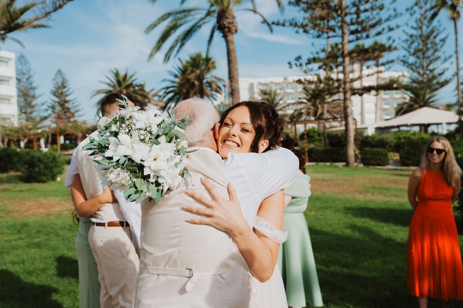 Two people in light colored attire share a joyful embrace outdoors in a sunny garden setting with palm trees.
