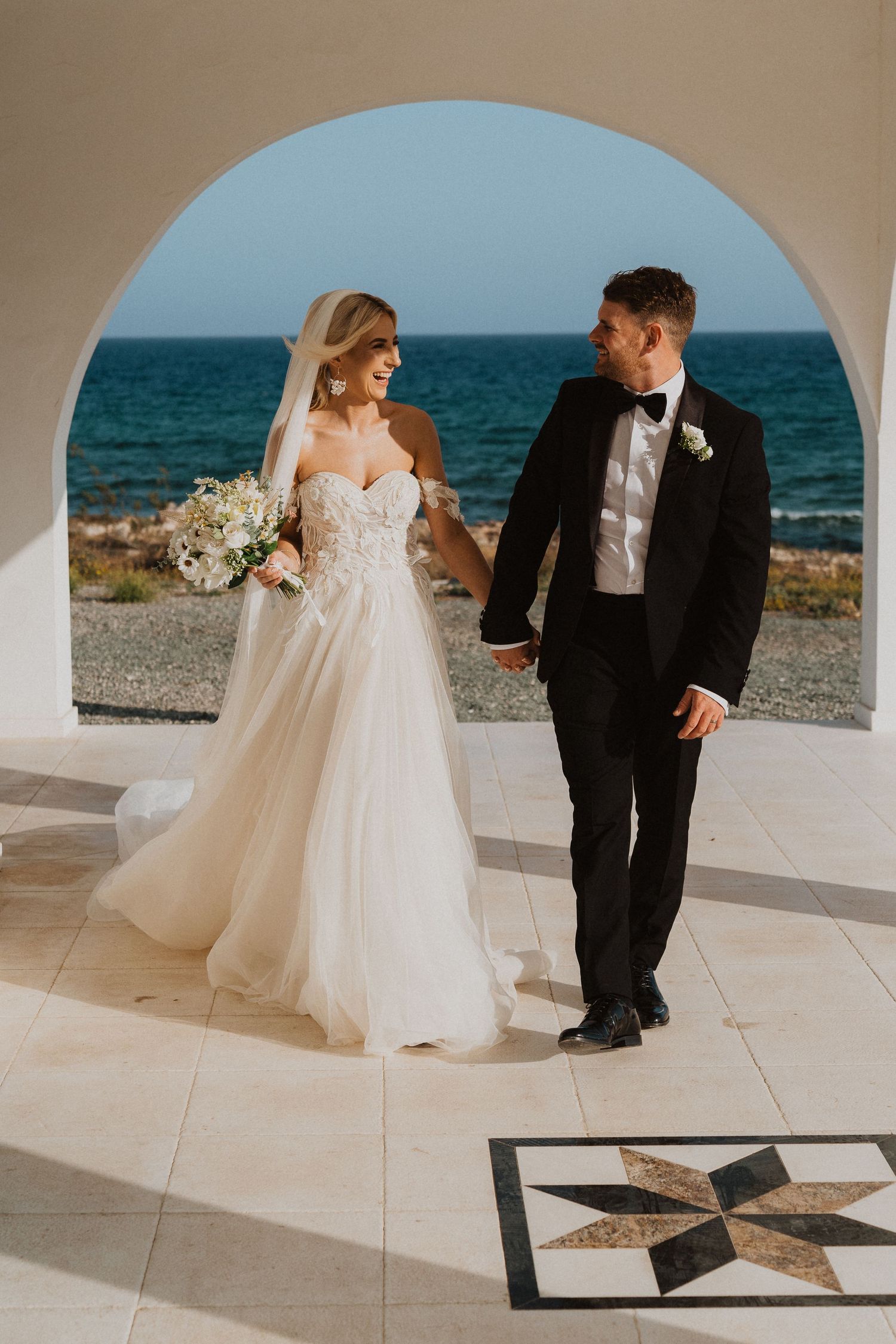 Bride and groom walking through white arched doorway with ocean view and tiled floor.