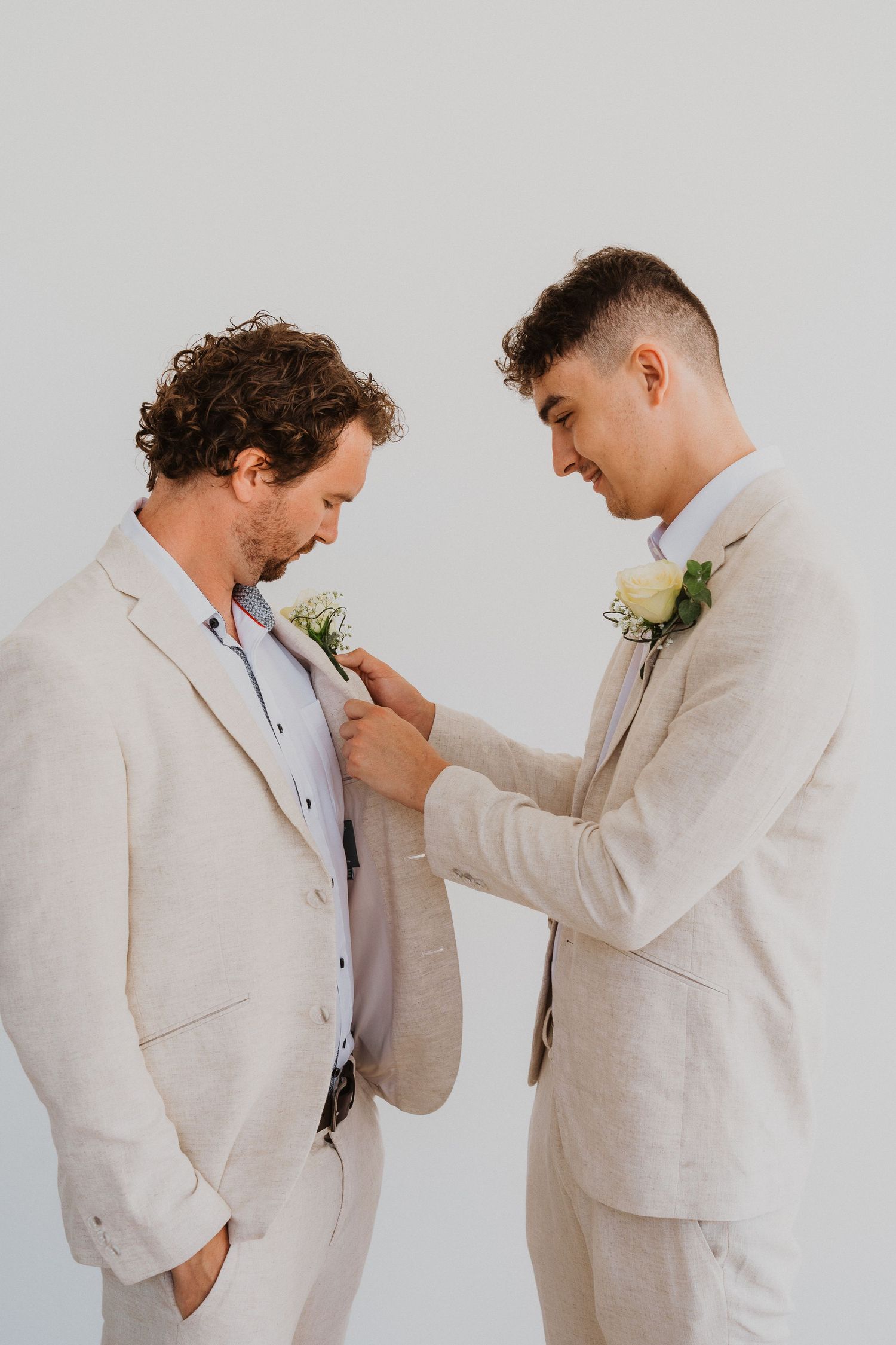 Two groomsmen in beige suits share a touching moment as one adjusts the other's boutonniere before the ceremony.