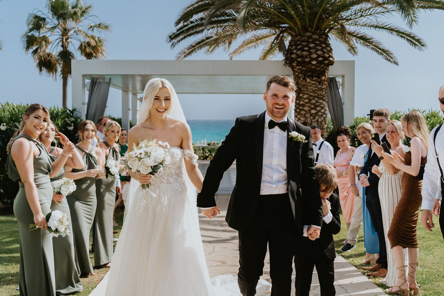 A wedding ceremony on a green lawn with palm trees and ocean views as guests line the aisle in celebration.