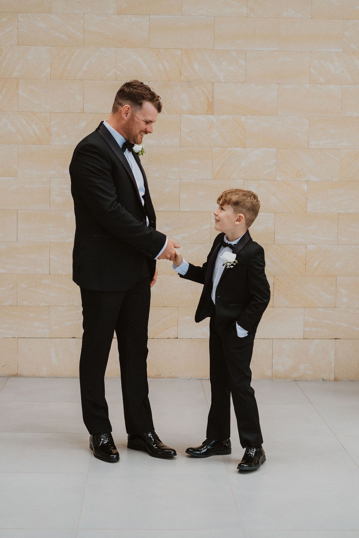 Two people in matching tuxedos having a playful moment against stone wall.