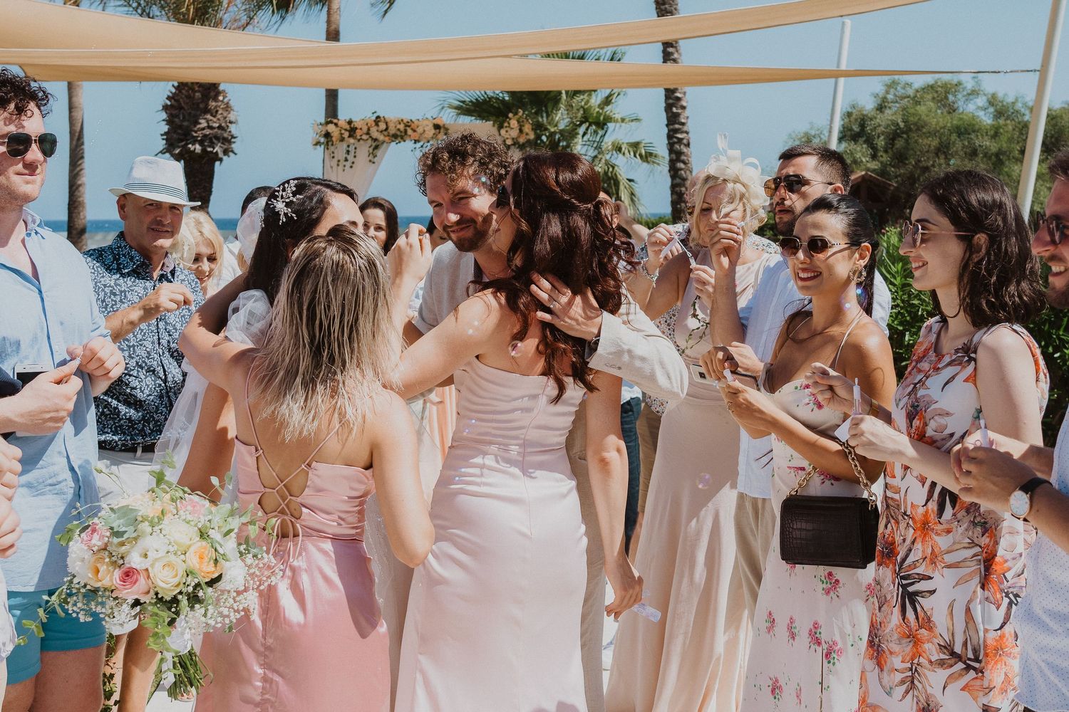 Wedding party celebrates joyfully under beige canopy at outdoor beach venue.