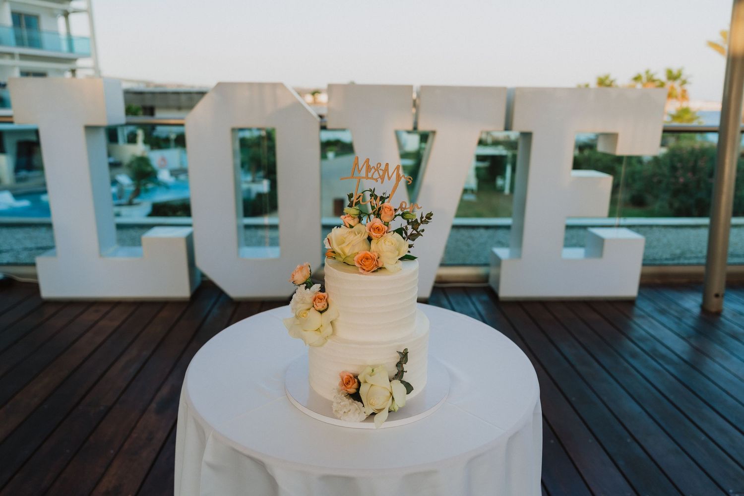 Wedding cake adorned with peach and white flowers sits on a round table with white linens.