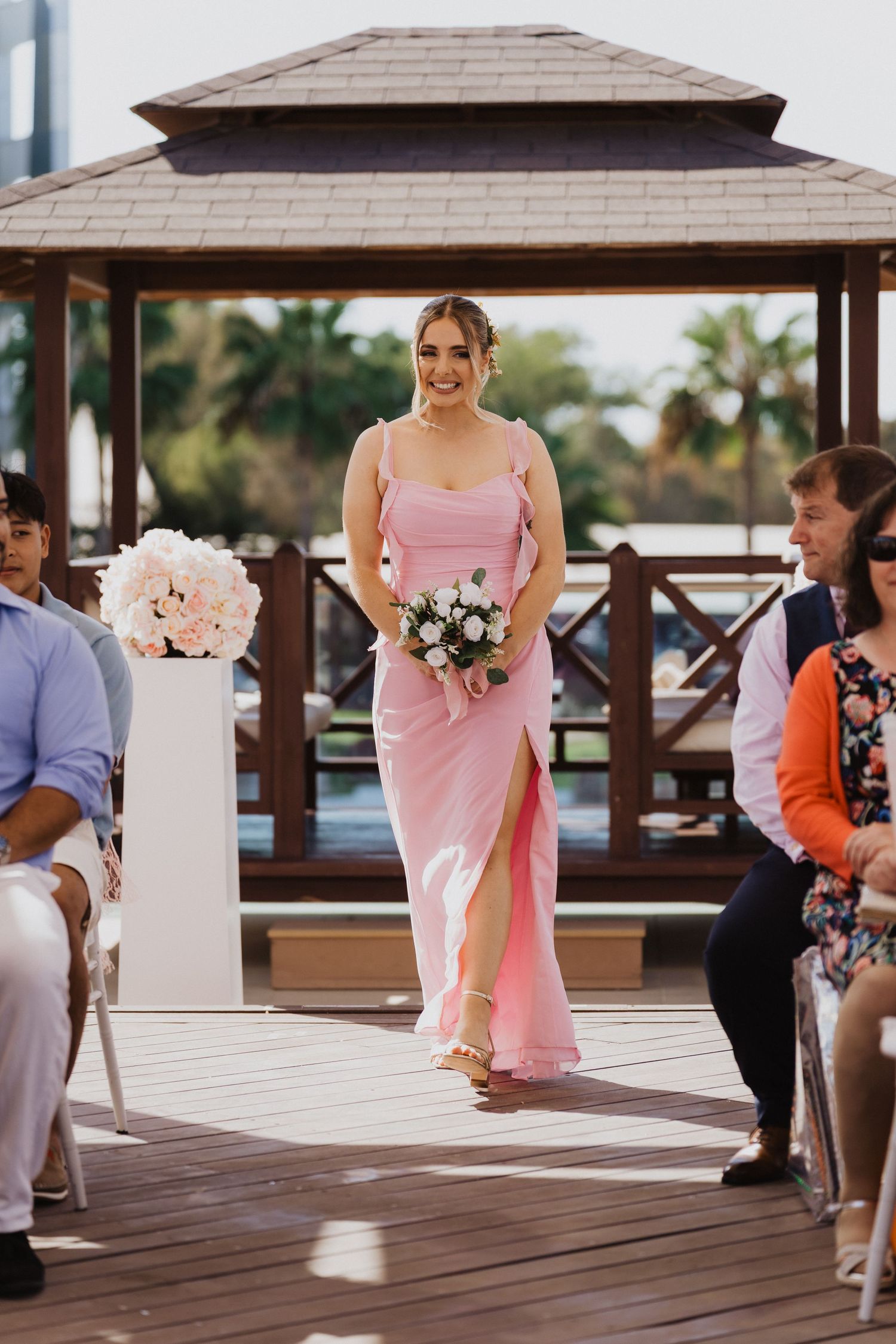 Bridesmaids in pink dresses process down a wooden deck aisle at a waterfront wedding venue.