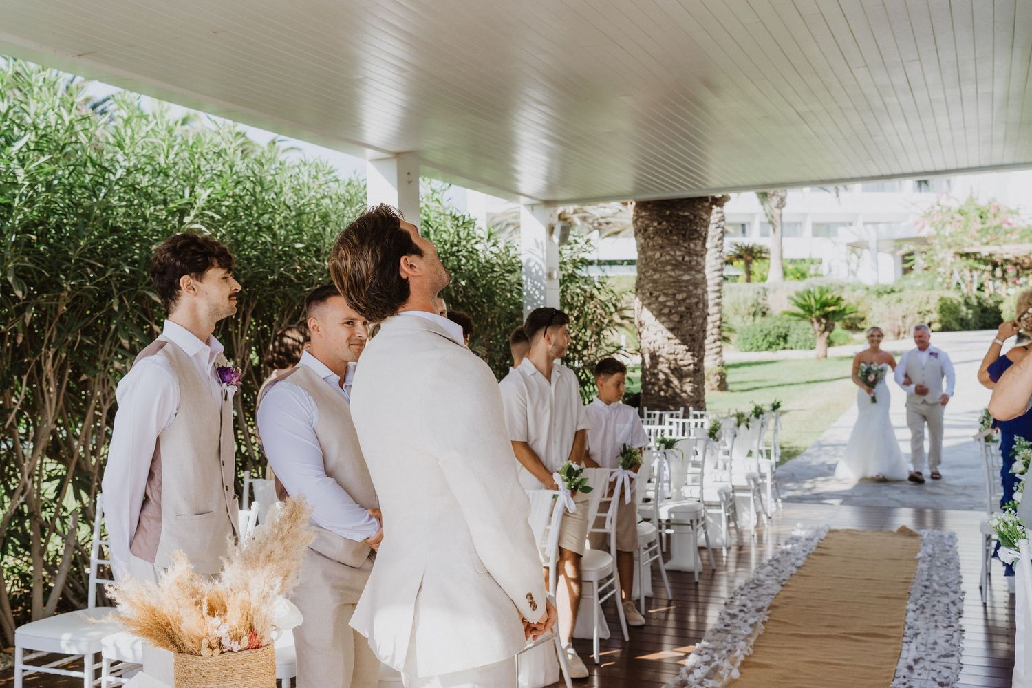 Wedding party members walk down a covered outdoor aisle decorated with white petals at a garden venue.
