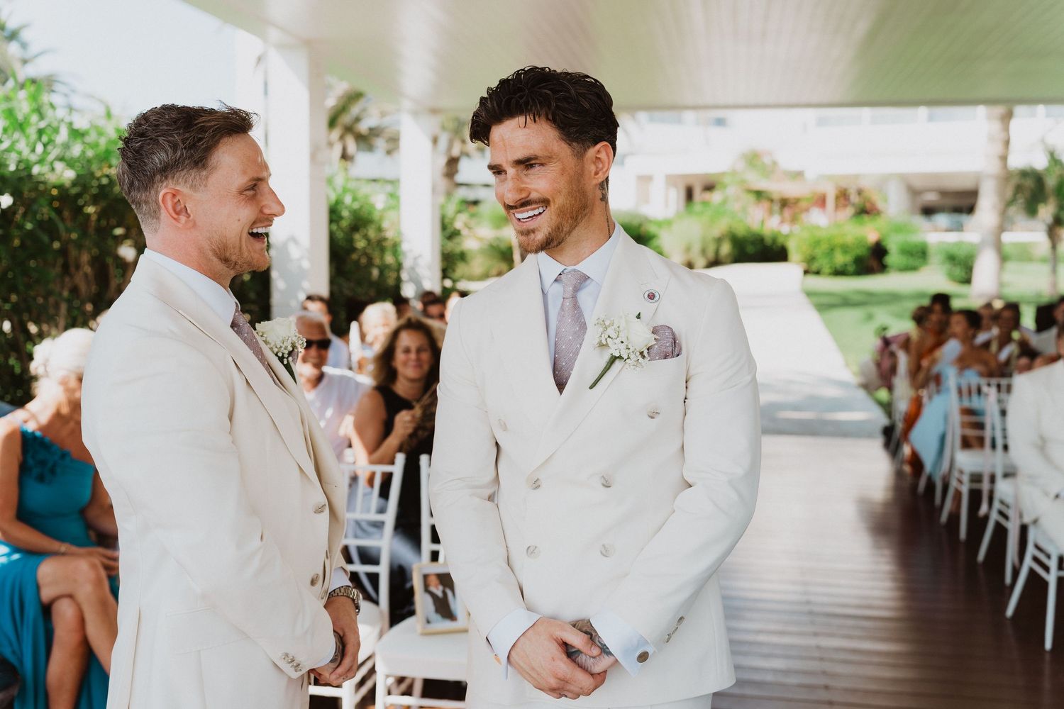 Two people in matching white suits share a joyful moment during an outdoor wedding ceremony.