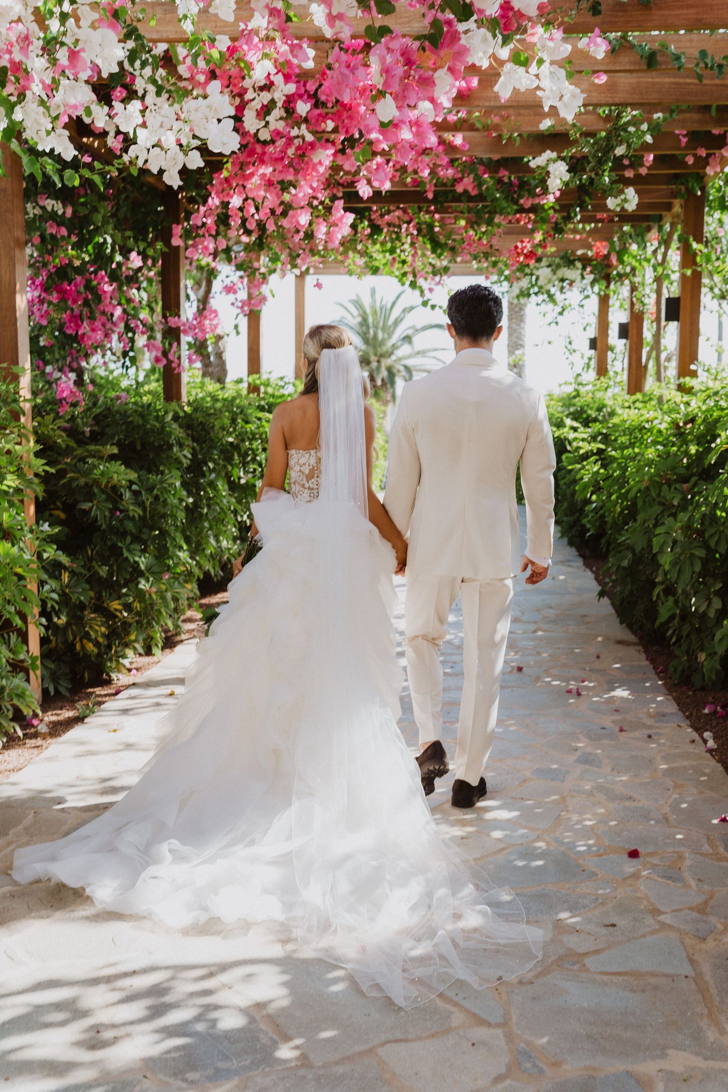 A couple walks down a garden pathway covered by pink bougainvillea flowers and wooden pergola on their wedding day.