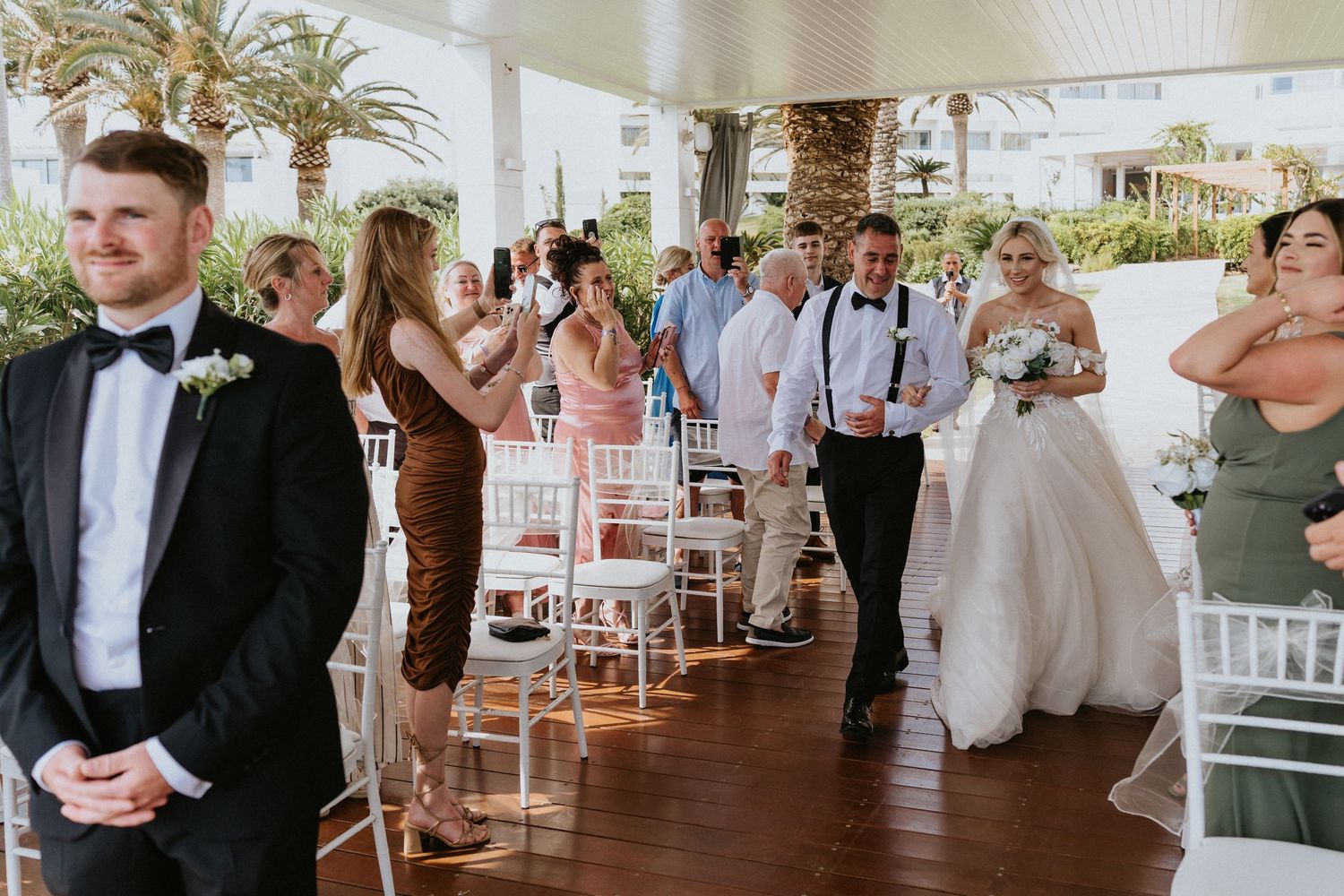 Wedding guests gathered on a covered patio with white chairs and tropical palm trees.