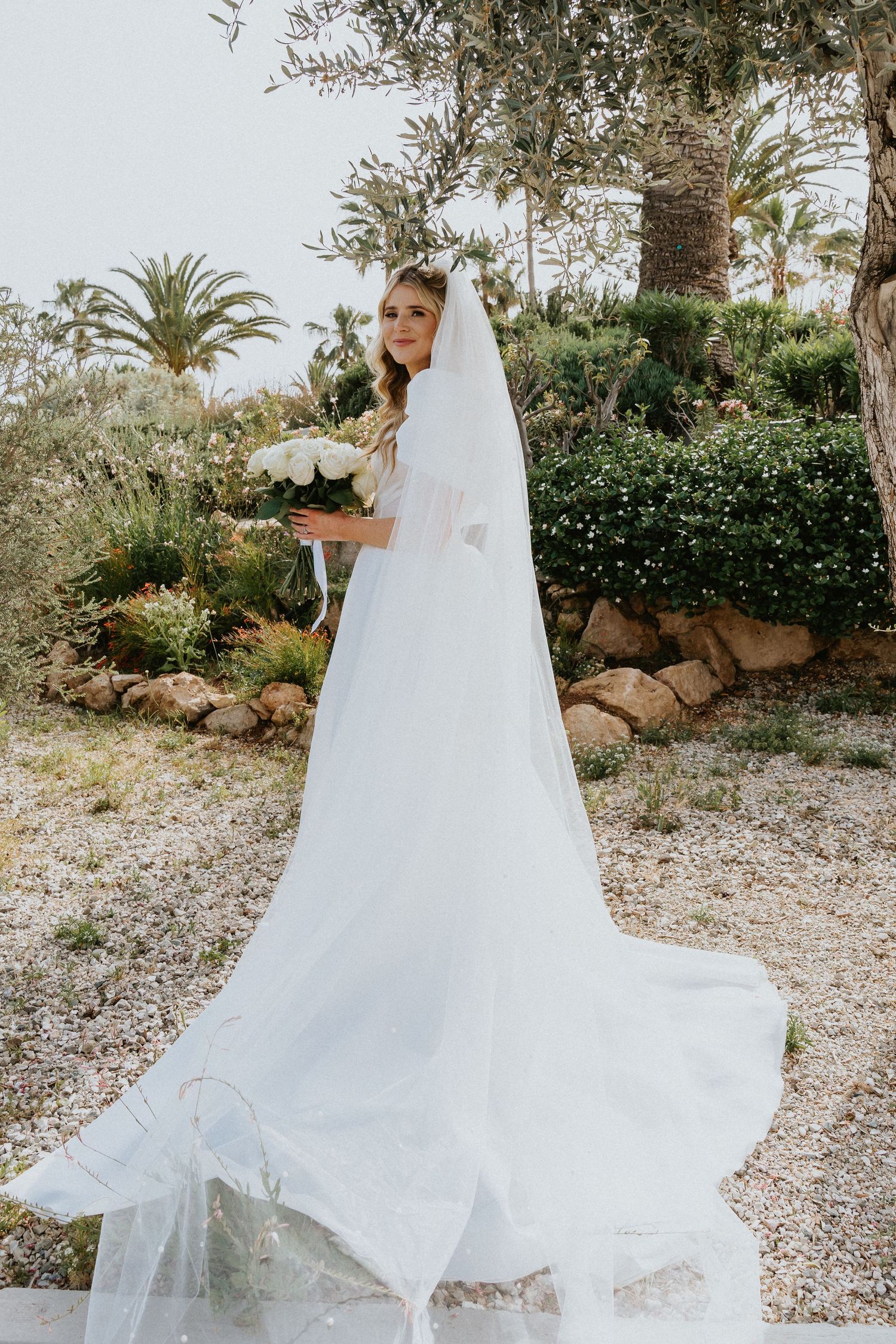 Bride in flowing white dress and veil poses with bouquet in tropical garden setting.