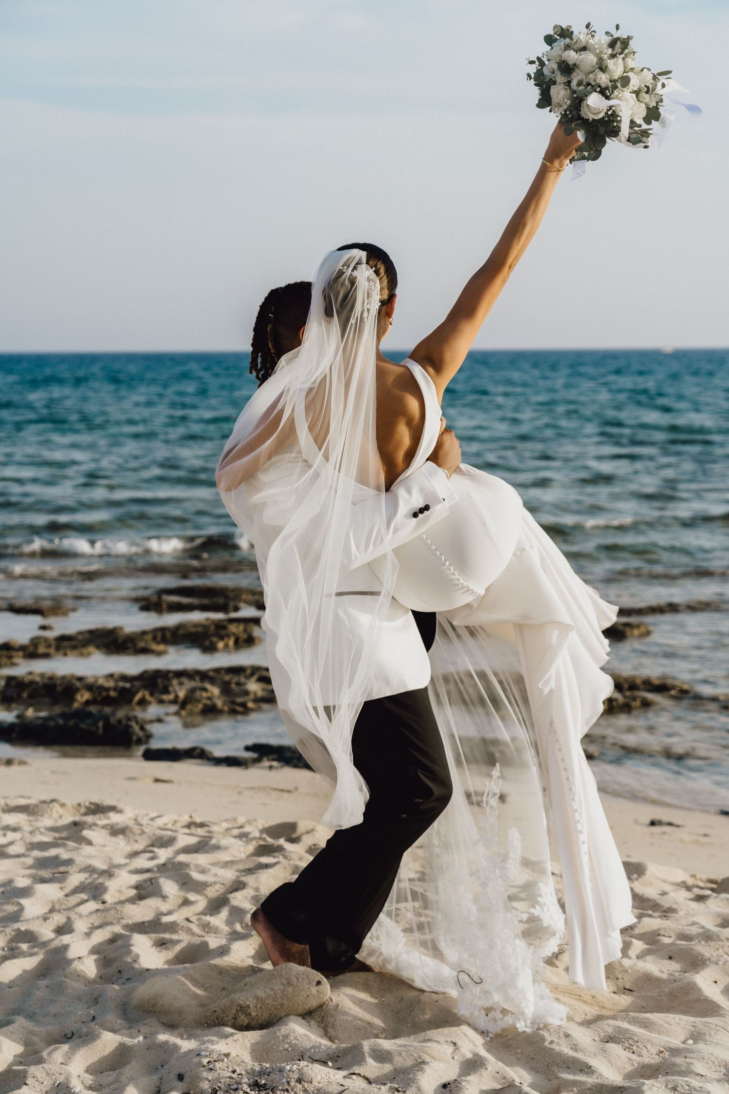 Couple in wedding attire playfully poses on beach with raised bouquet.