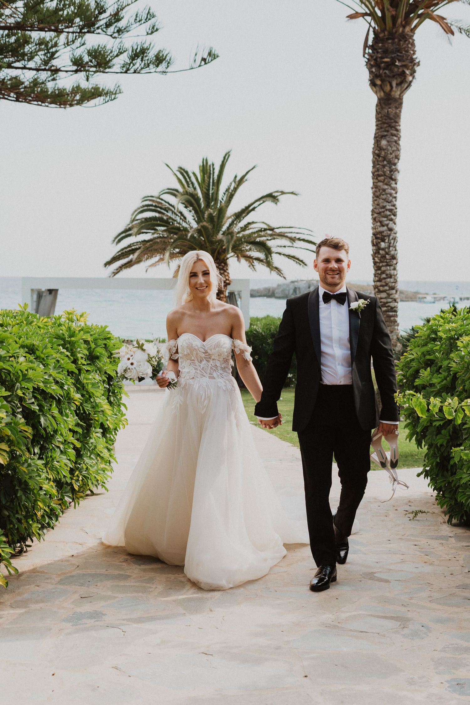 Wedding couple strolls along stone pathway surrounded by lush gardens and palm trees.