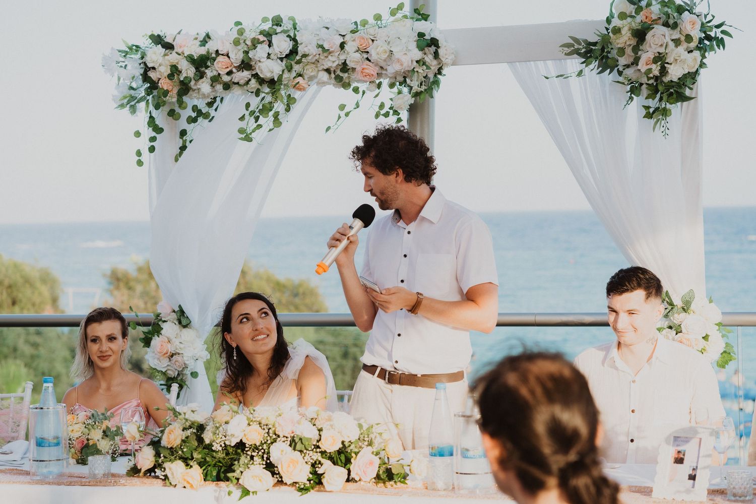 Speech time during seaside wedding reception under white canopy with floral decorations.