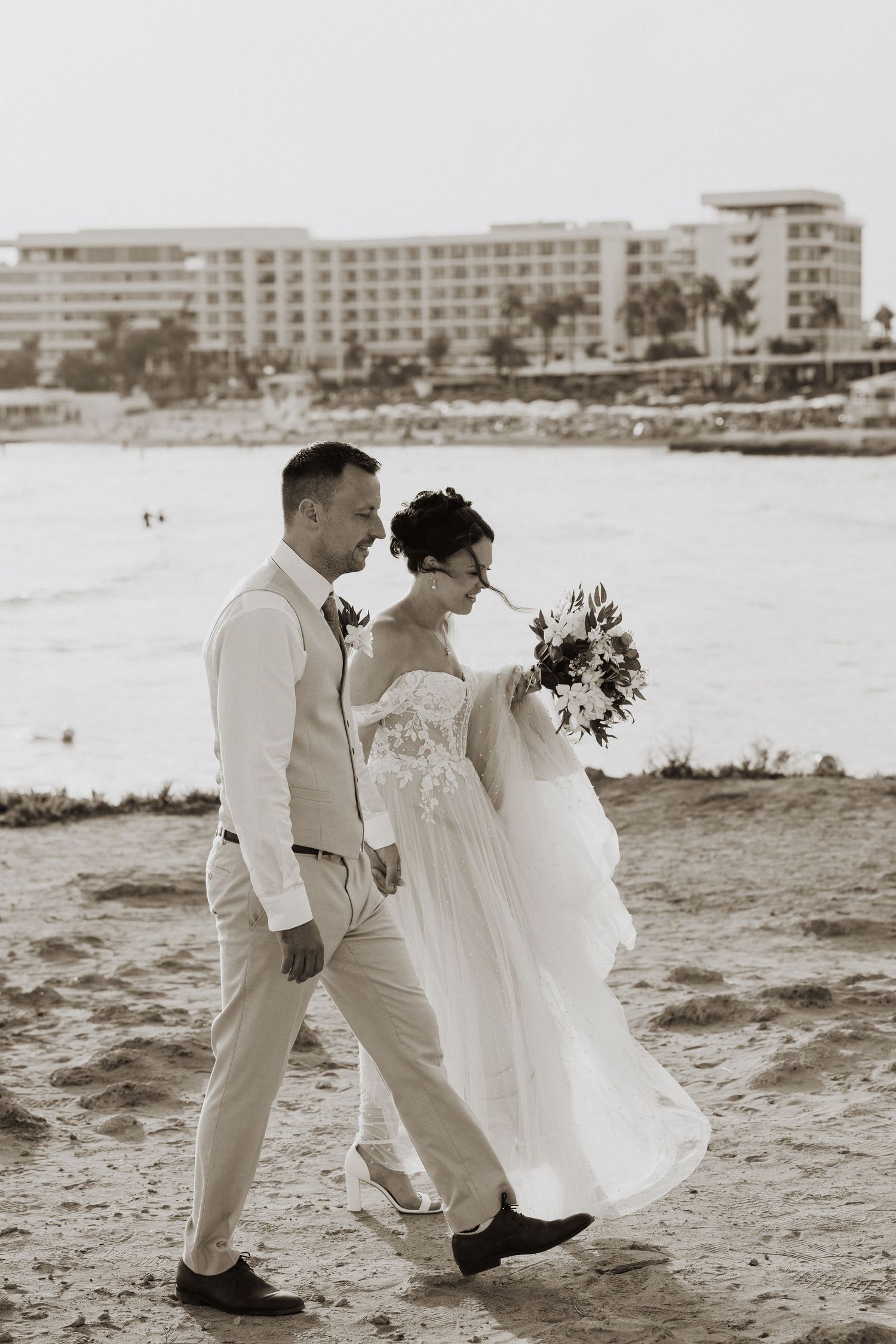 A couple in wedding attire walks along a beach with a resort hotel in the background in black and white photography.