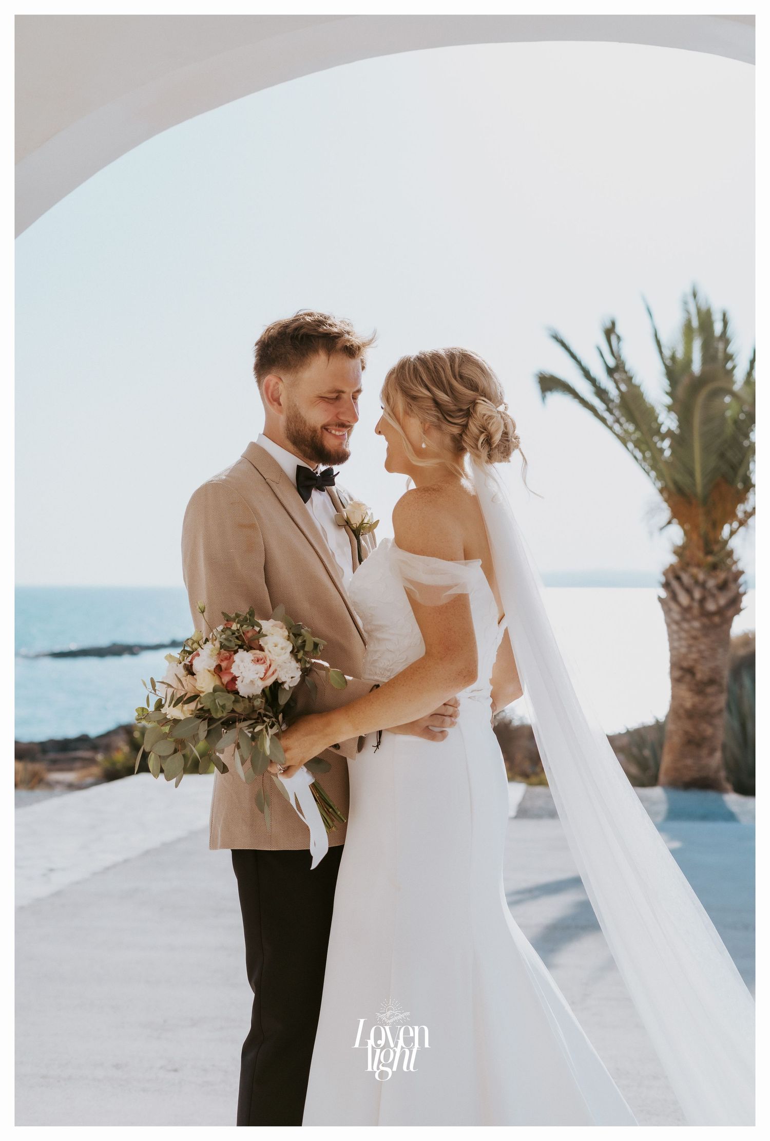 A romantic wedding photo with a couple embracing near palm trees and ocean views in a beachside setting.