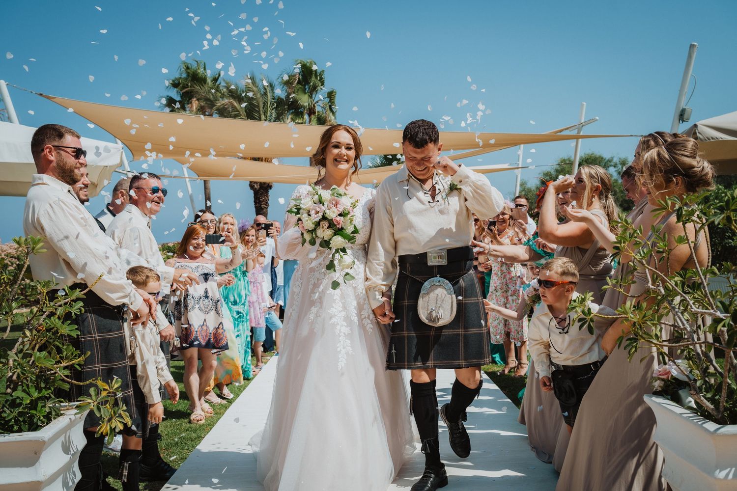 Happy wedding couple walks through confetti shower on outdoor aisle with tropical backdrop.