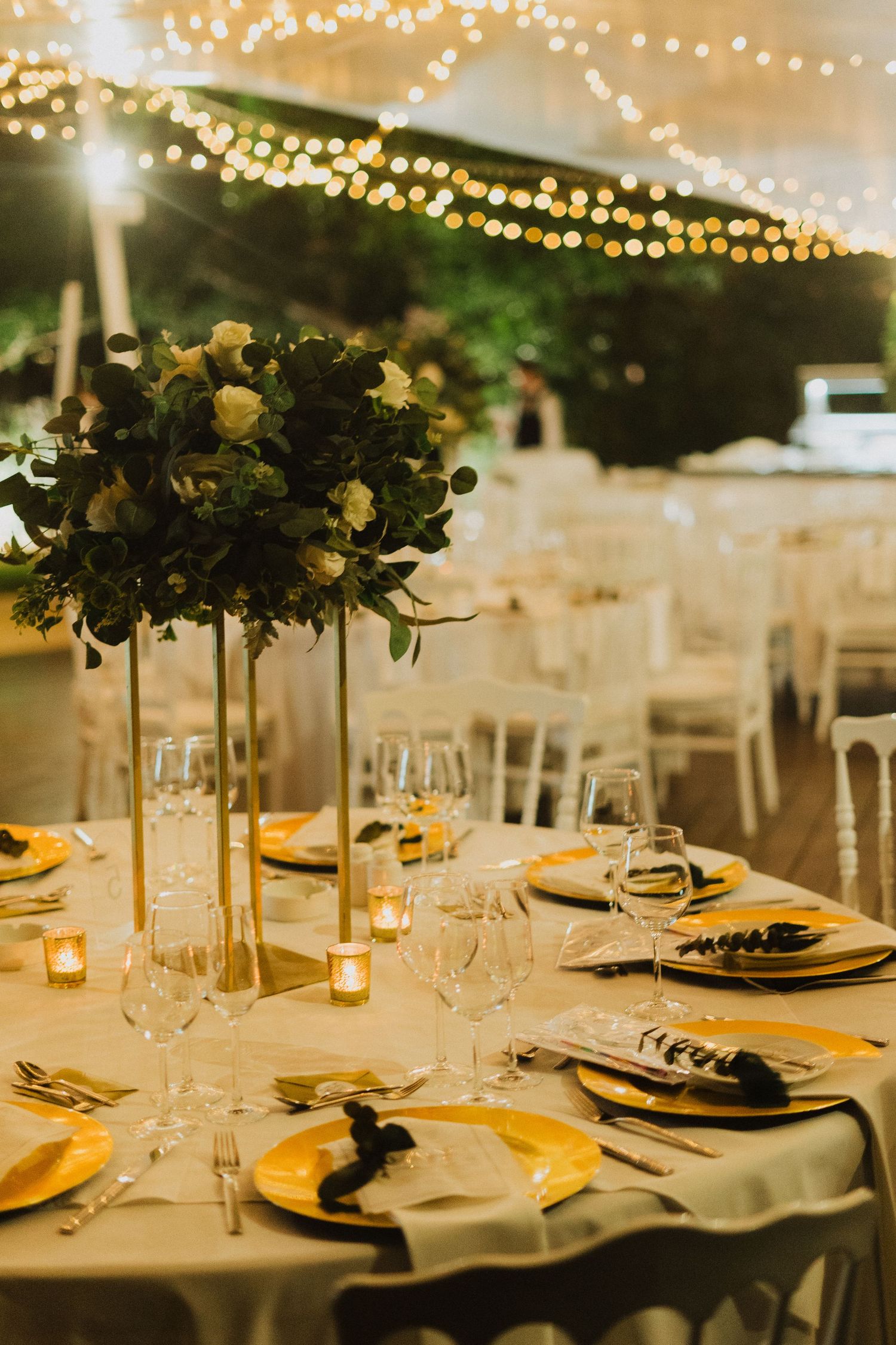 Close-up of a wedding table setting with tall floral centerpieces and gold accents under twinkling lights.