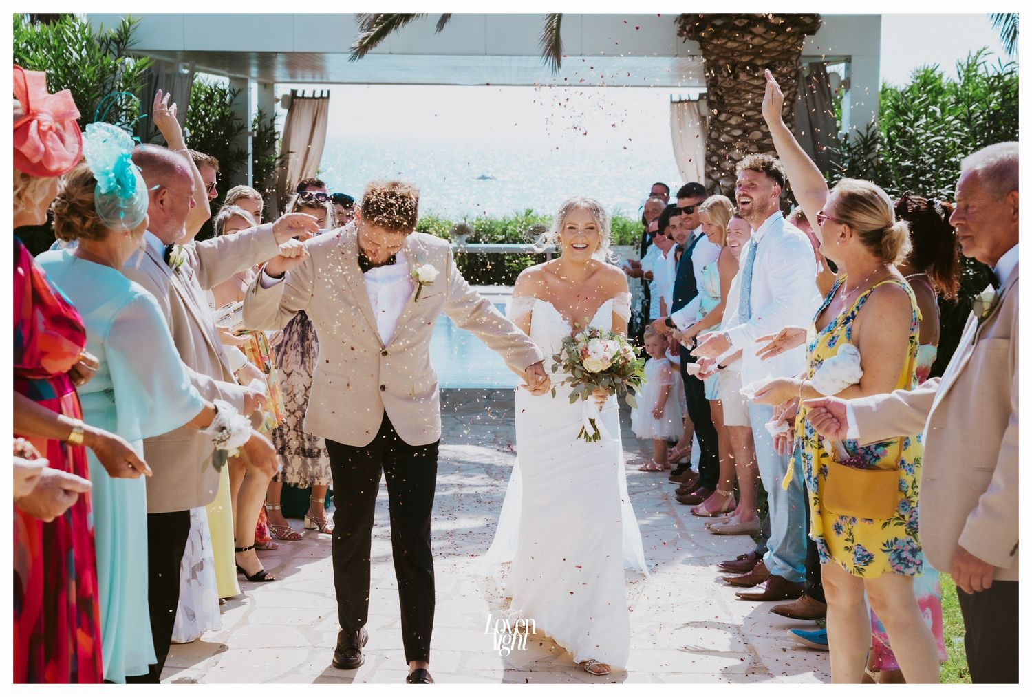 Guests throw confetti as newlyweds walk down an outdoor aisle at a tropical venue.