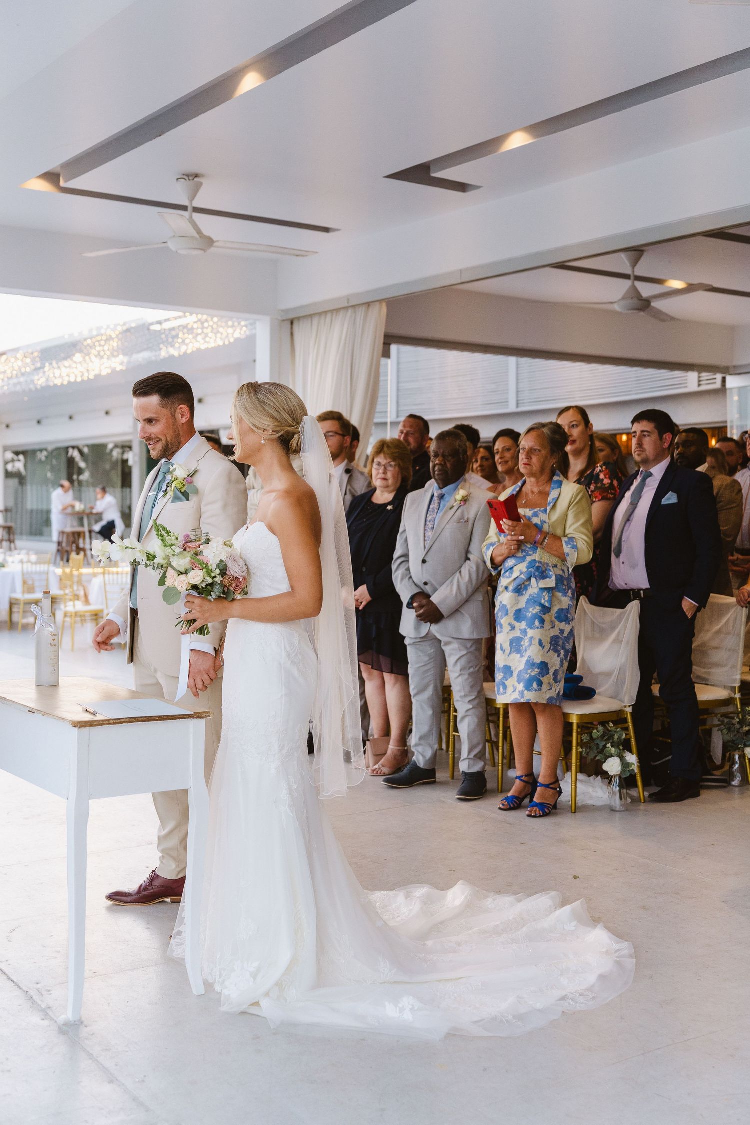 Modern wedding ceremony in bright venue with standing guests and white floral decorations.