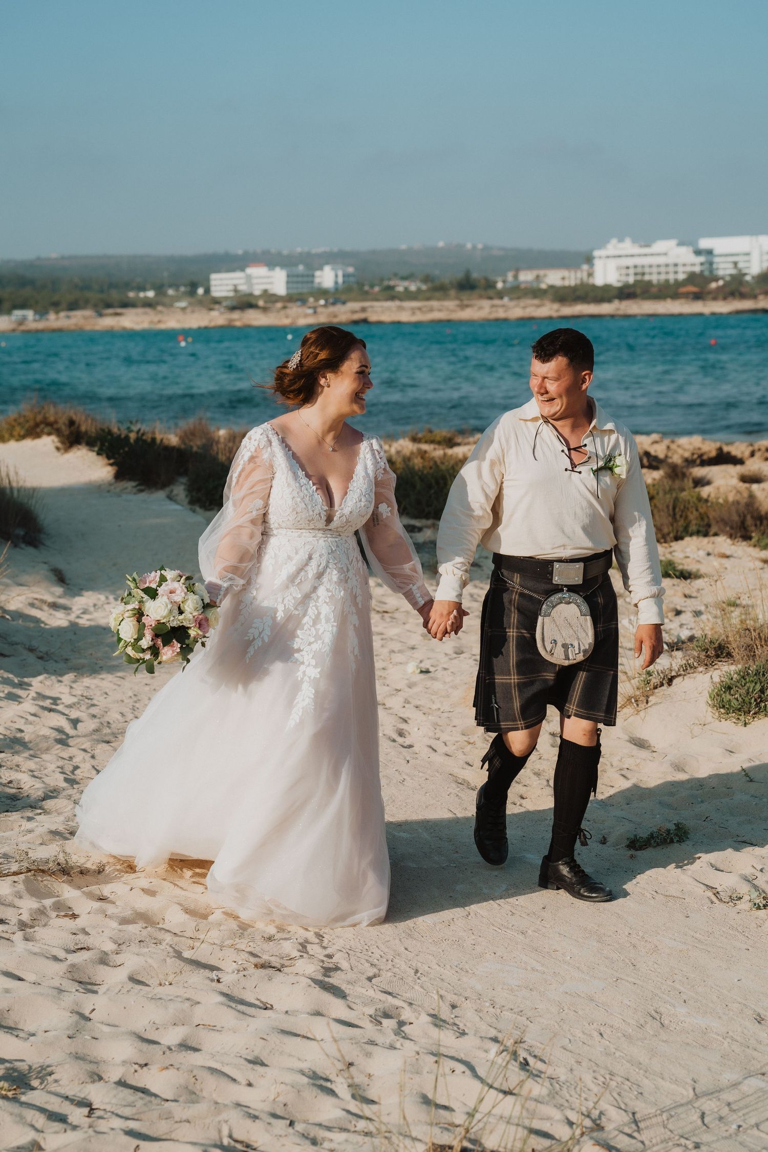 A romantic beach stroll with white wedding dress and traditional Scottish kilt against azure waters.
