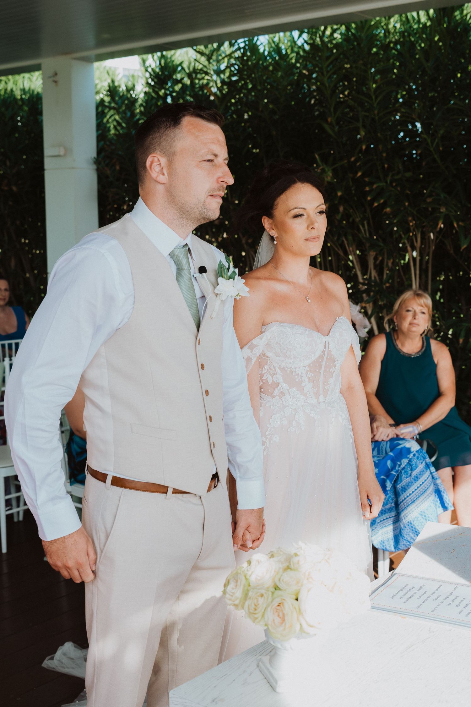 A couple stands together during their wedding ceremony, with the bride in white lace and groom in light beige suit.