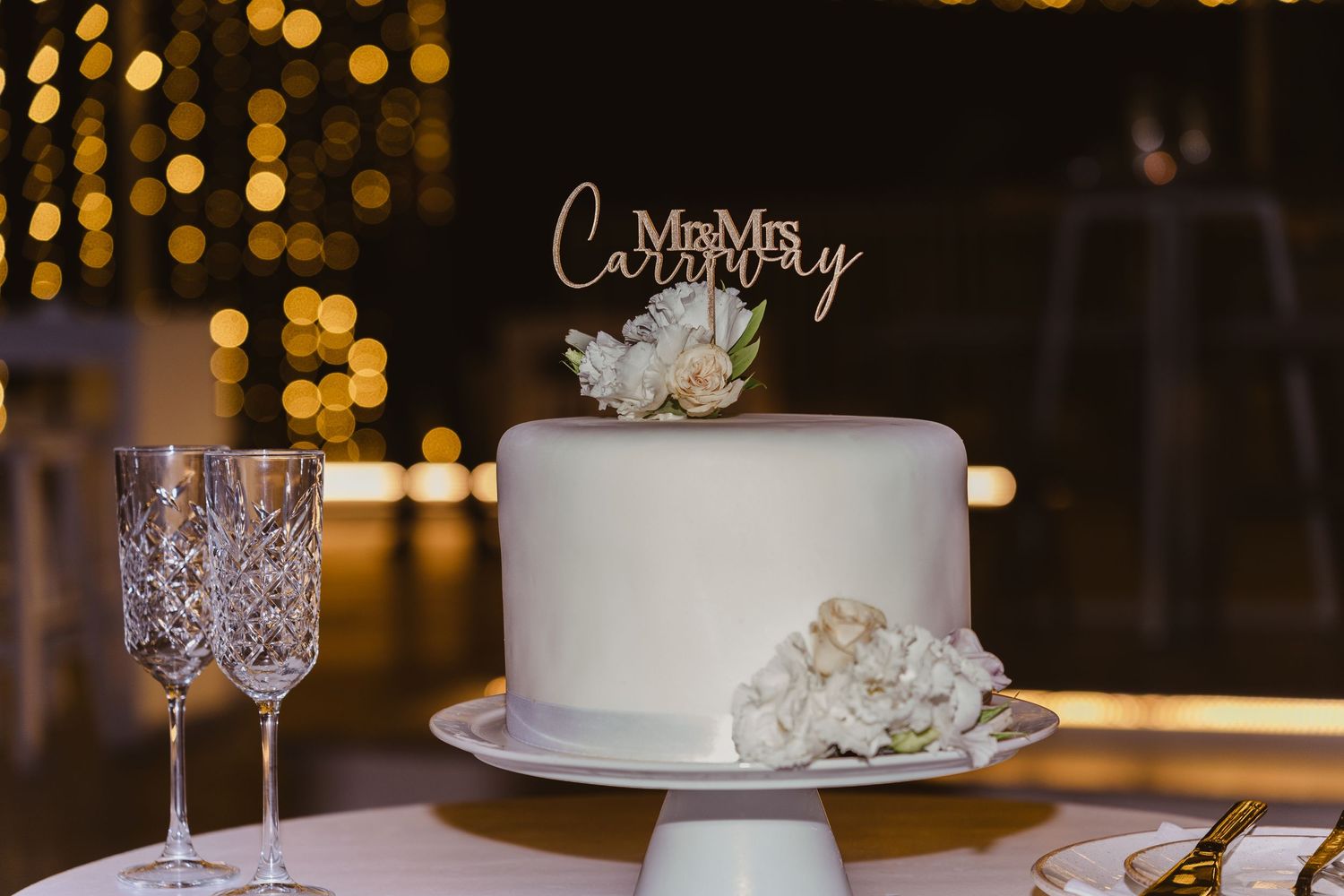 White wedding cake with 'Mr & Mrs' topper and floral decorations displayed against bokeh lights.