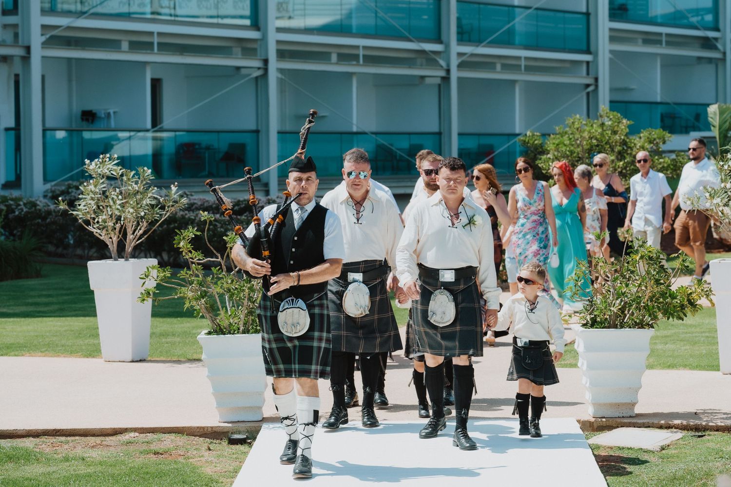 Group in matching kilts leads wedding procession down outdoor walkway.