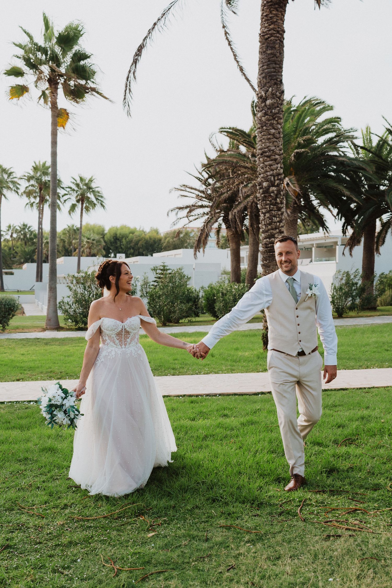 Happy couple in wedding attire walk hand in hand on grass with tropical palm trees in background at outdoor venue.