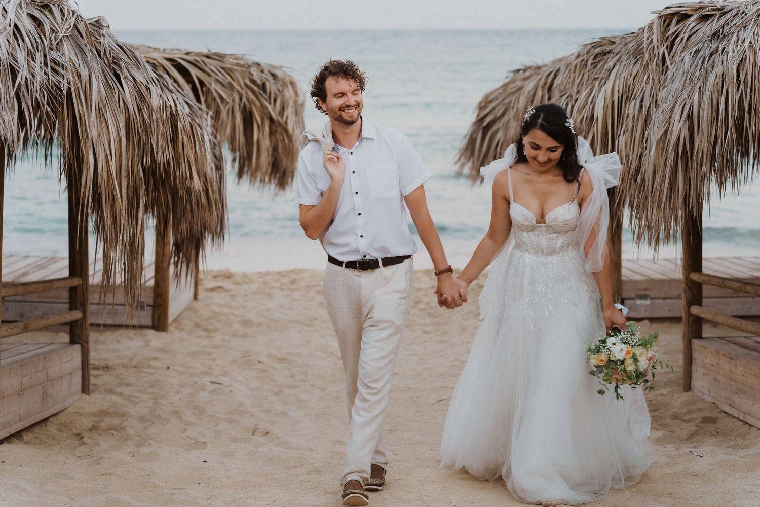 A joyful couple walks hand in hand on a beach with thatched palapas in the background.