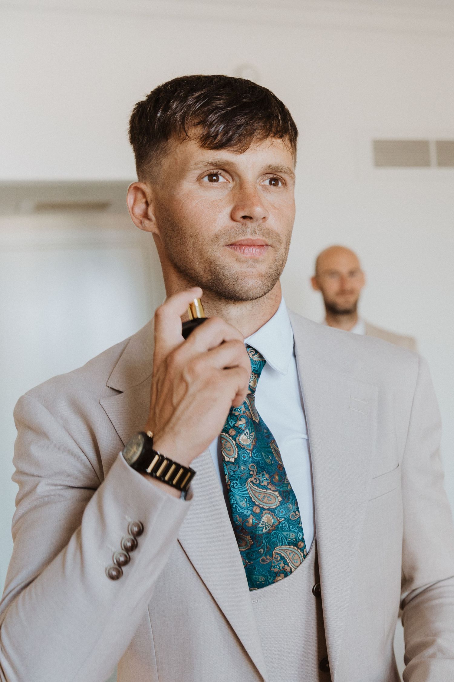 A person in a light gray suit adjusts their teal patterned tie while another person stands in the background.