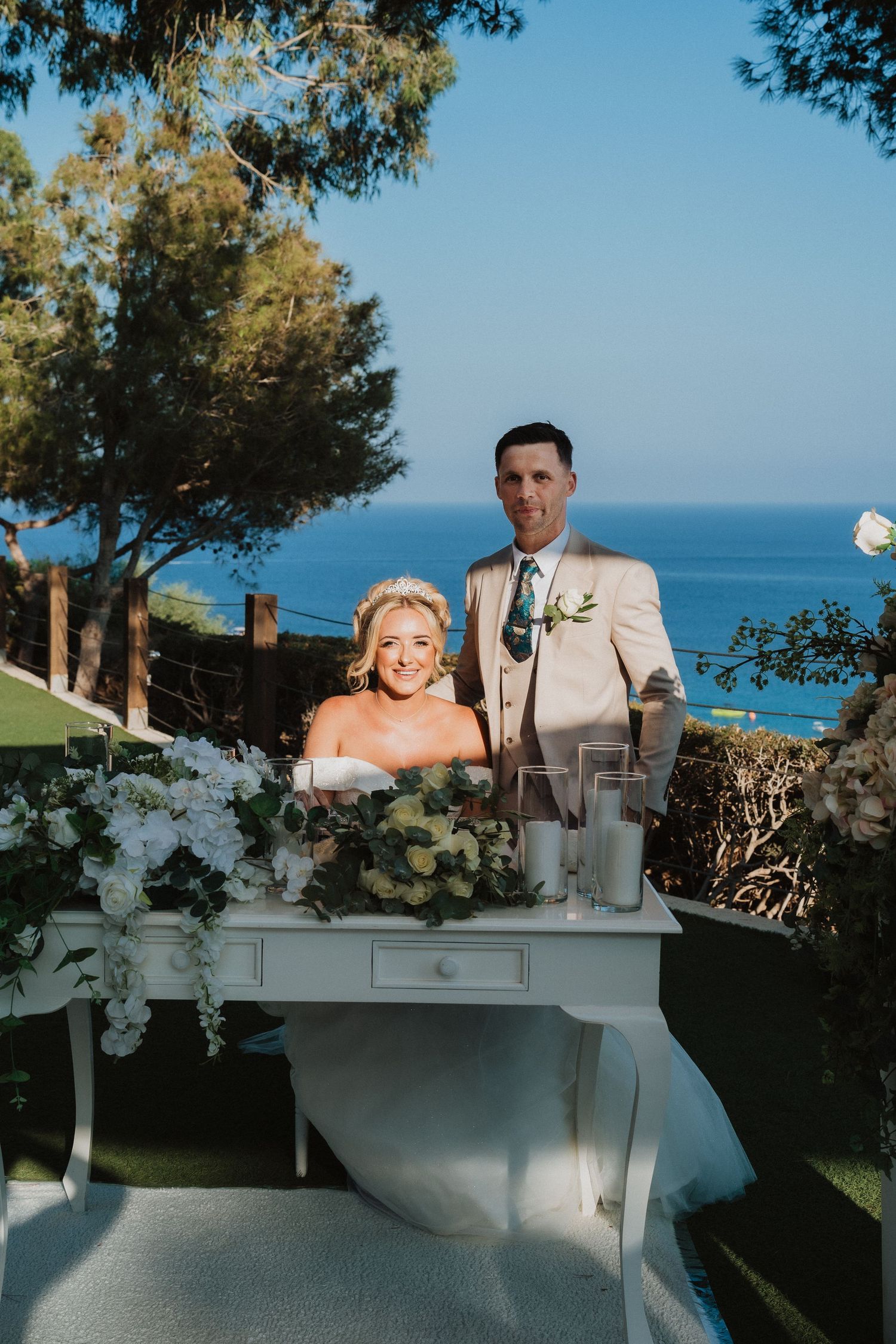 Seaside wedding reception with an elegant white desk adorned with floral arrangements overlooking the ocean.