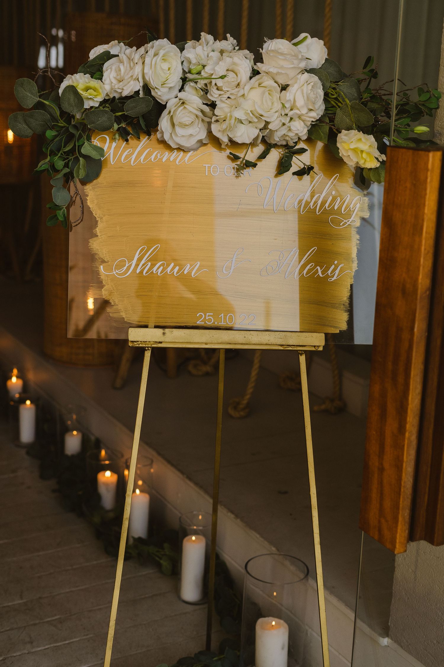 An elegant wedding welcome sign decorated with white roses and lit by glowing candles.
