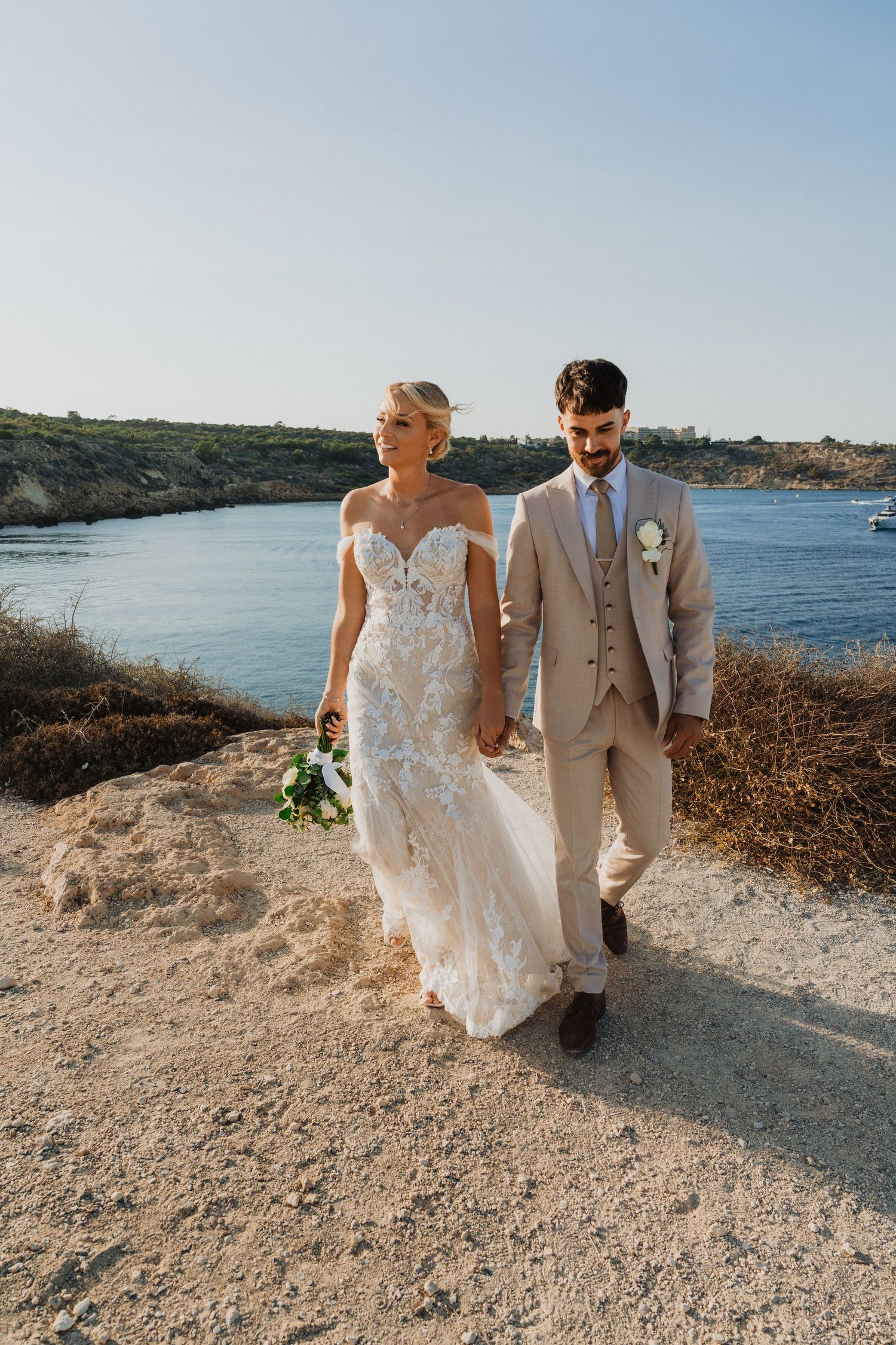 A romantic coastal wedding photoshoot featuring a couple walking along a rocky Mediterranean path.