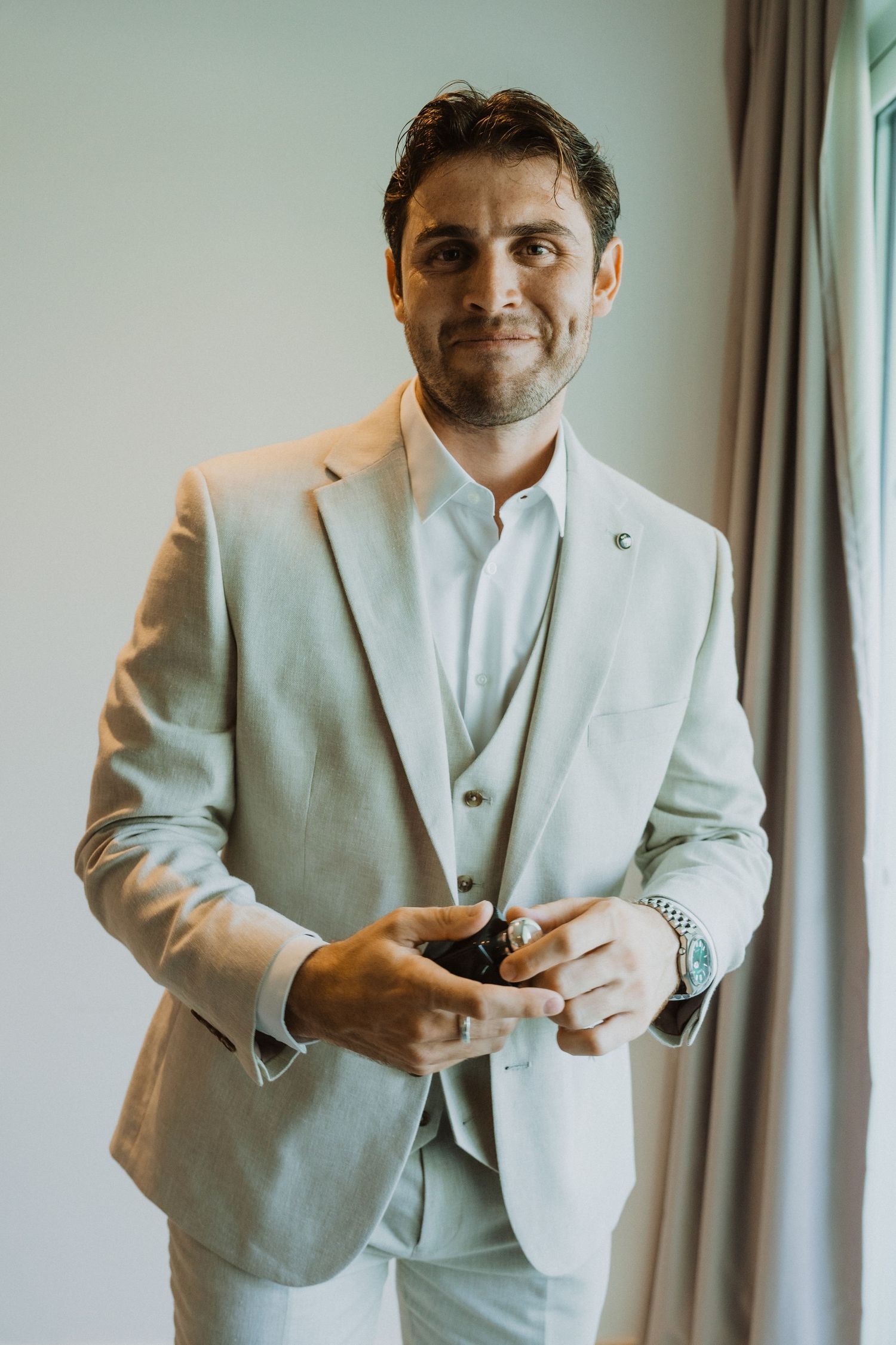 A groom in a light gray three-piece suit adjusts his buttons while standing by a window.