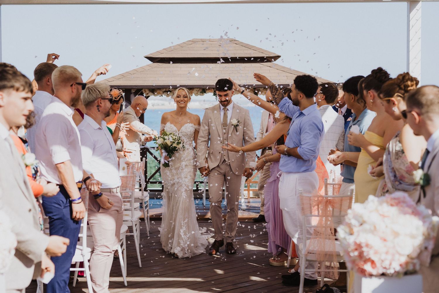 Wedding guests celebrate with confetti on an outdoor terrace by the sea.