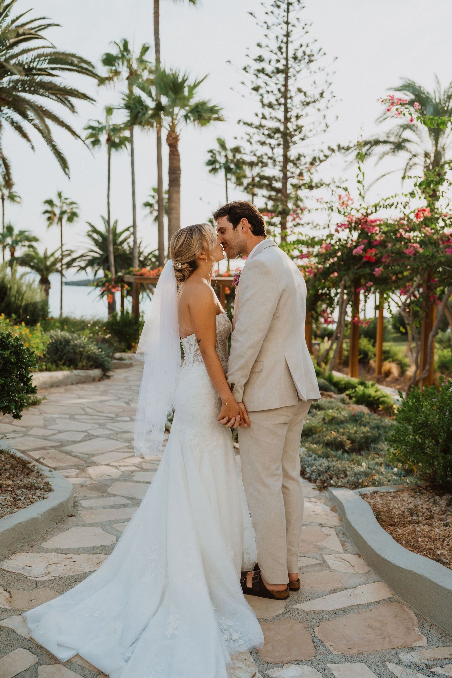 Newlyweds share a romantic moment on a stone pathway surrounded by tropical palm trees and flowers.