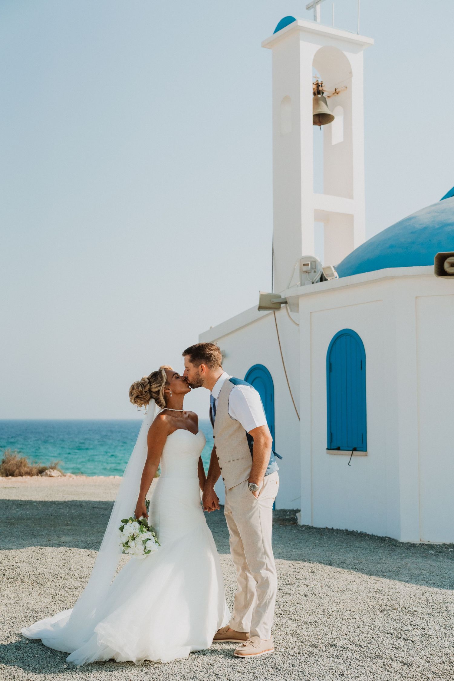 Bride and groom kissing in front of a white Greek chapel with blue dome by the sea on their wedding day.