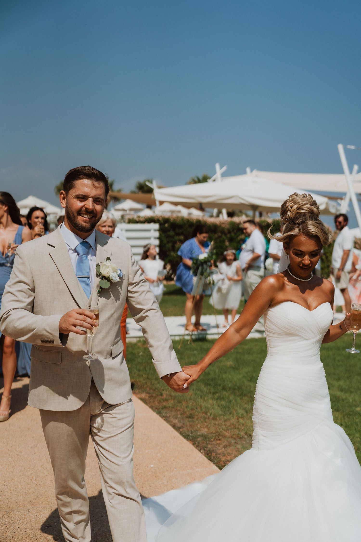 Smiling groom in tan suit holds champagne while walking hand-in-hand with bride in strapless white gown at outdoor wedding.