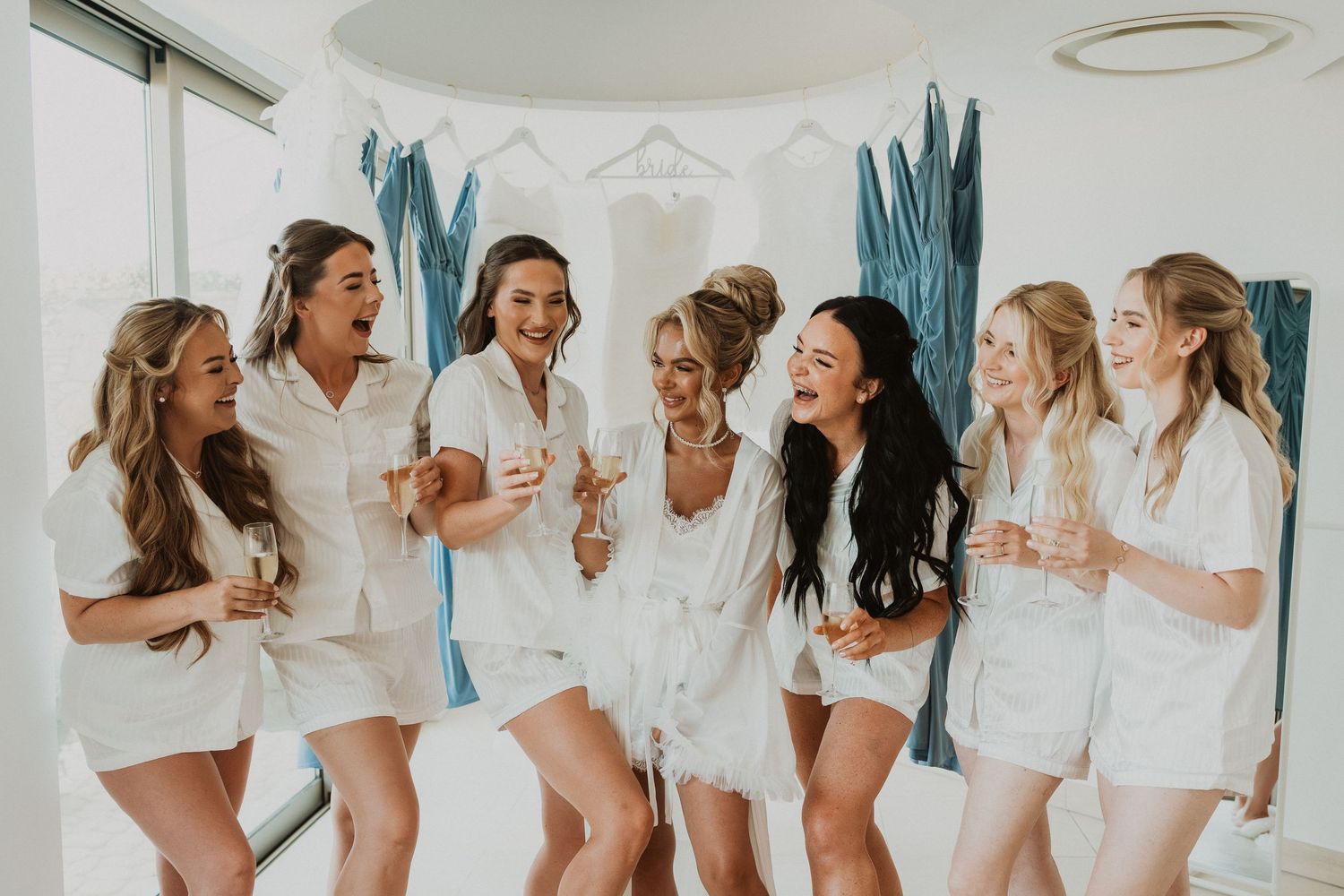Seven bridesmaids in white satin pajamas laughing and celebrating together during bridal party getting ready photos.