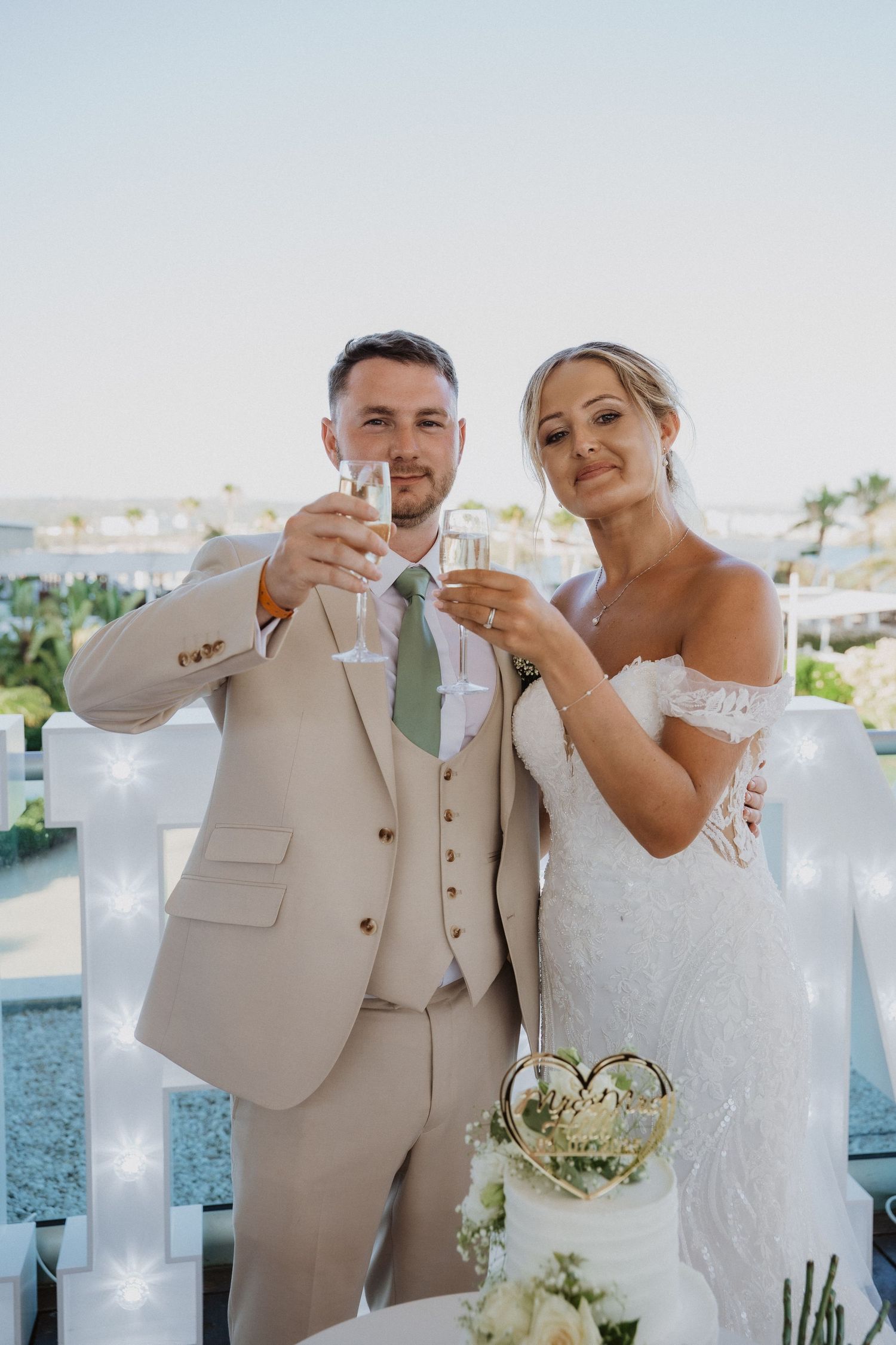 A couple in formal wedding attire toasts with champagne glasses against a waterfront backdrop.