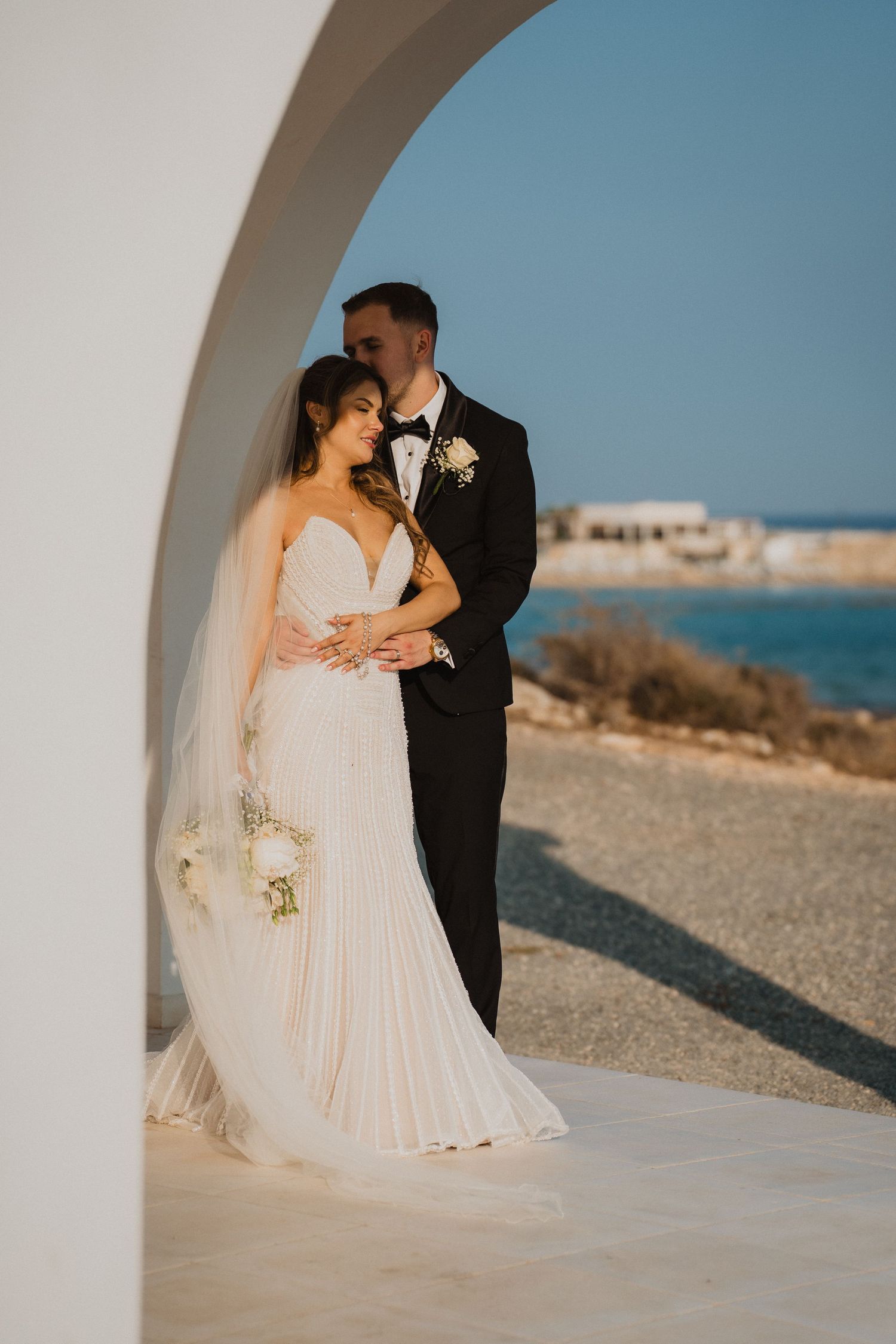 Bride in white gown and groom in tuxedo embrace under a white arch with ocean backdrop.