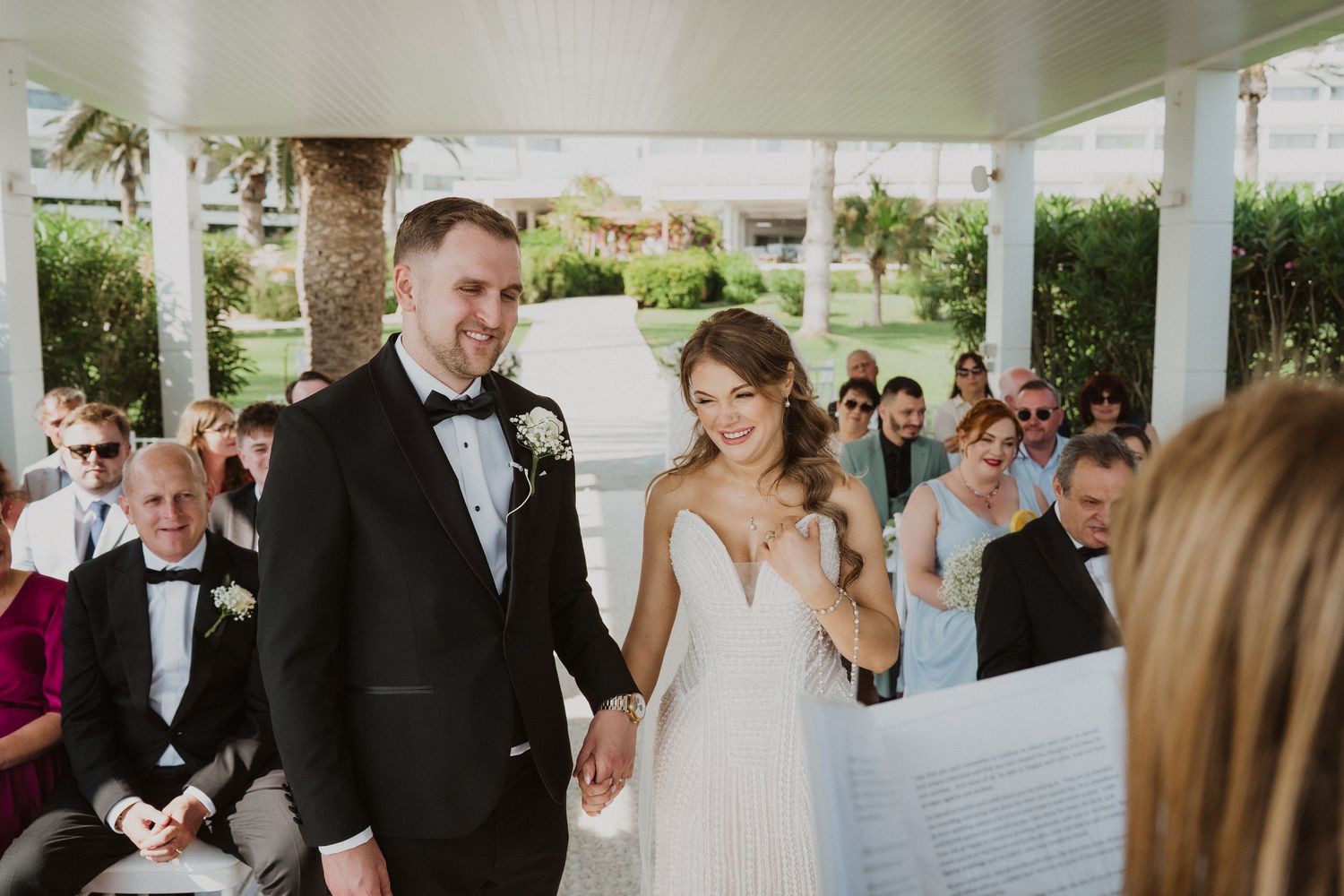 Smiling bride and groom holding hands at outdoor wedding ceremony with guests seated behind them.