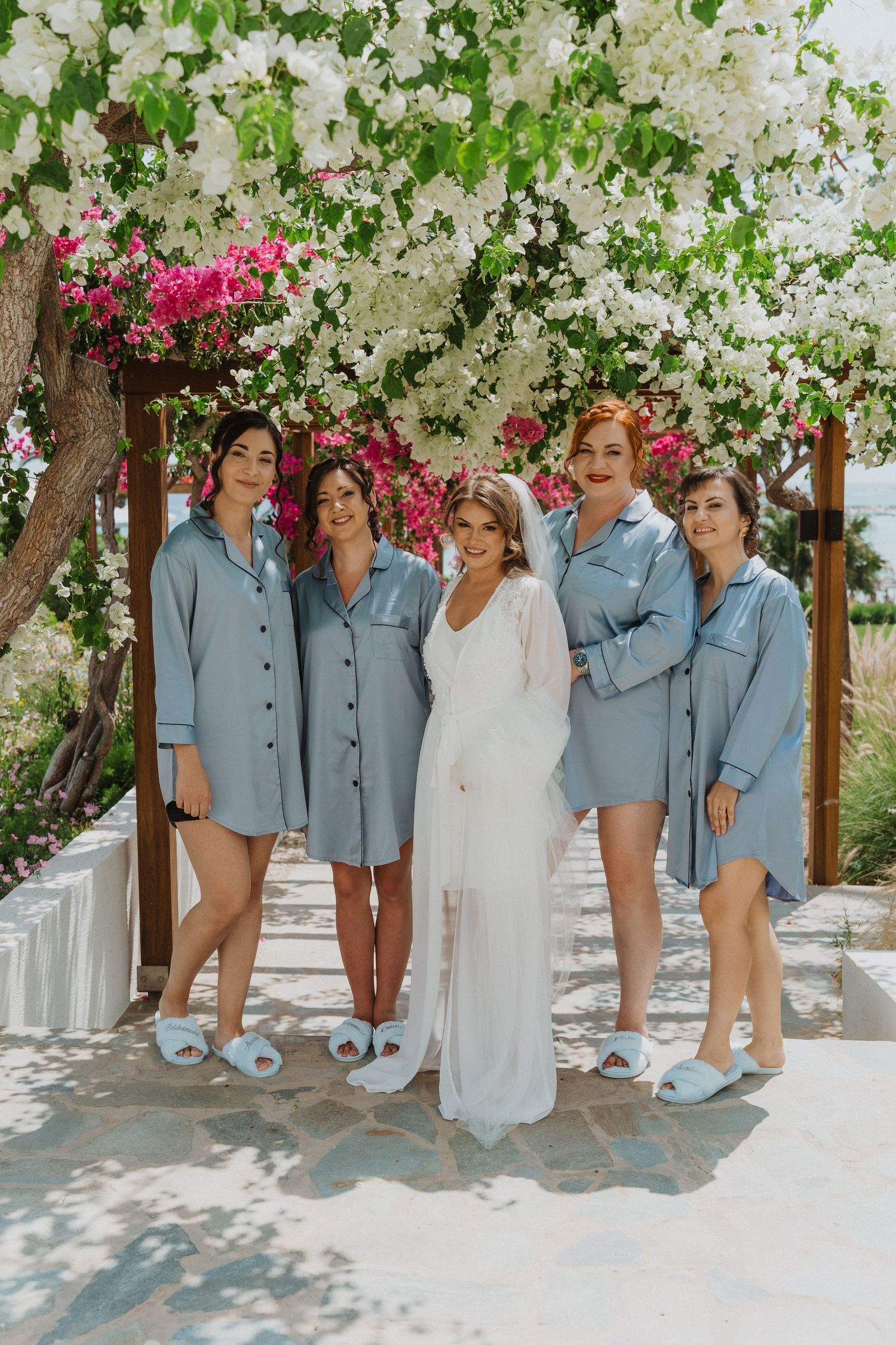 Bride in white gown poses with four bridesmaids in matching blue satin shirts and fuzzy slippers under blooming flowers.