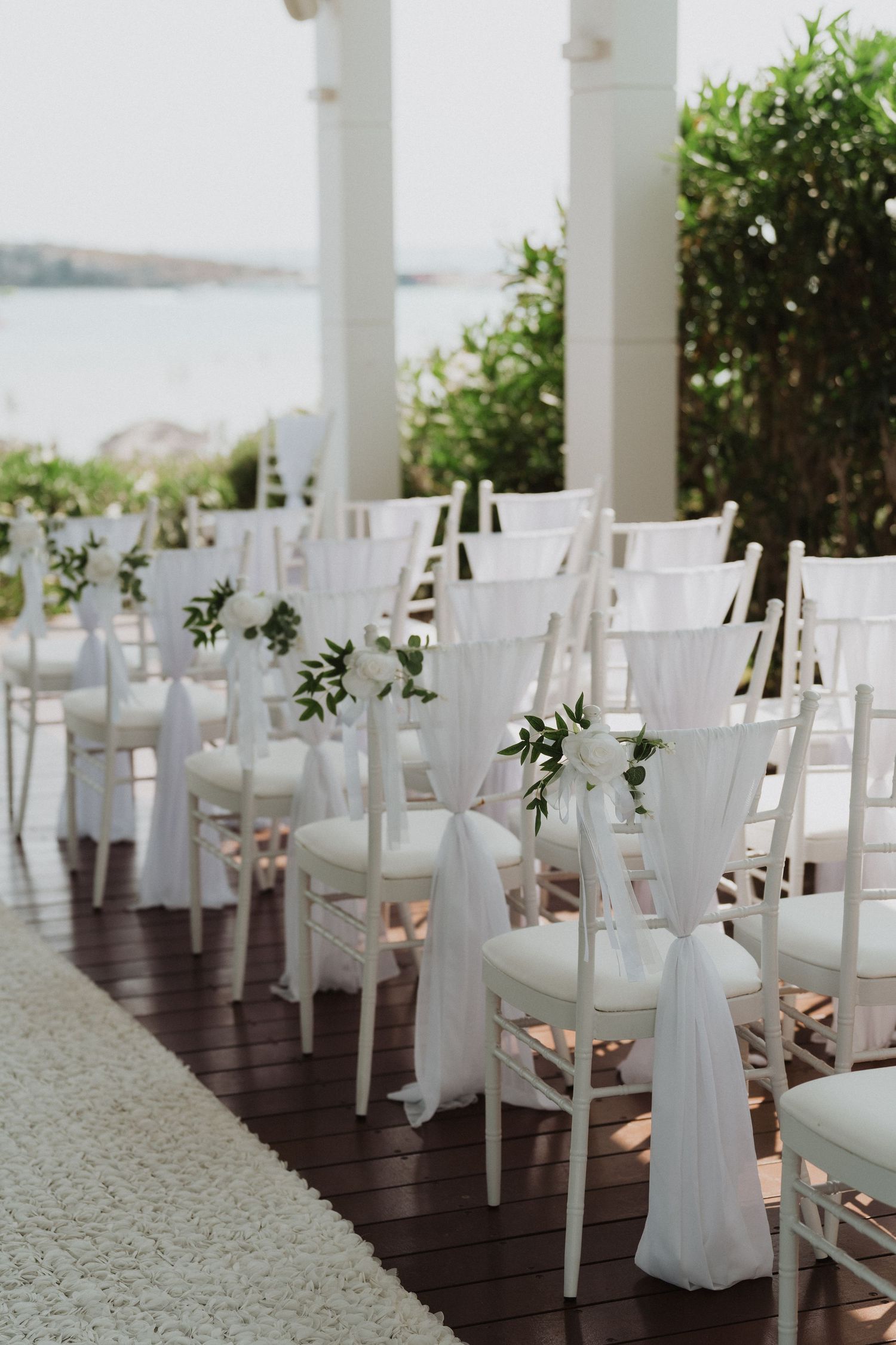 Elegant rows of white Chiavari chairs with white draping and greenery overlooking the sea.