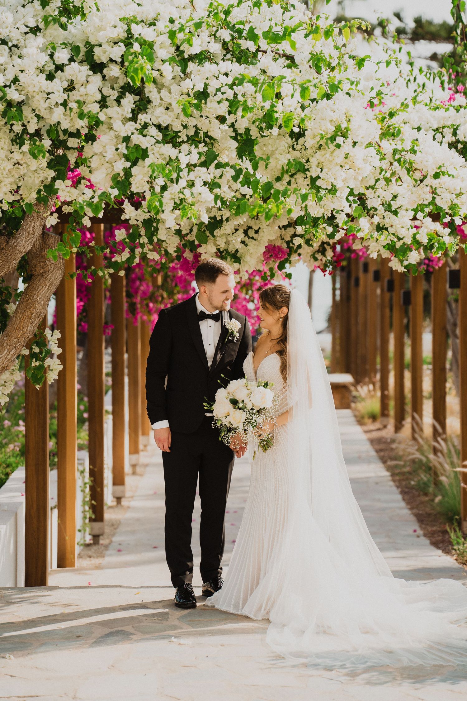 Wedding couple walks under a stunning white bougainvillea arbor with pink flowers in the background.