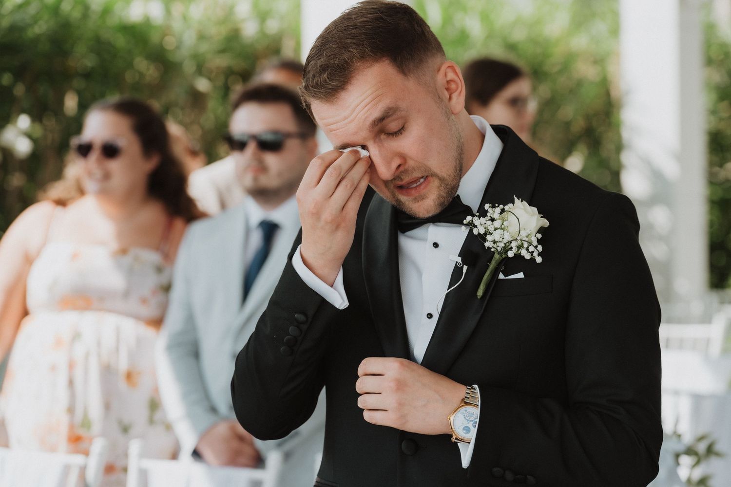 Emotional groom in black tuxedo wiping tears during outdoor wedding ceremony.