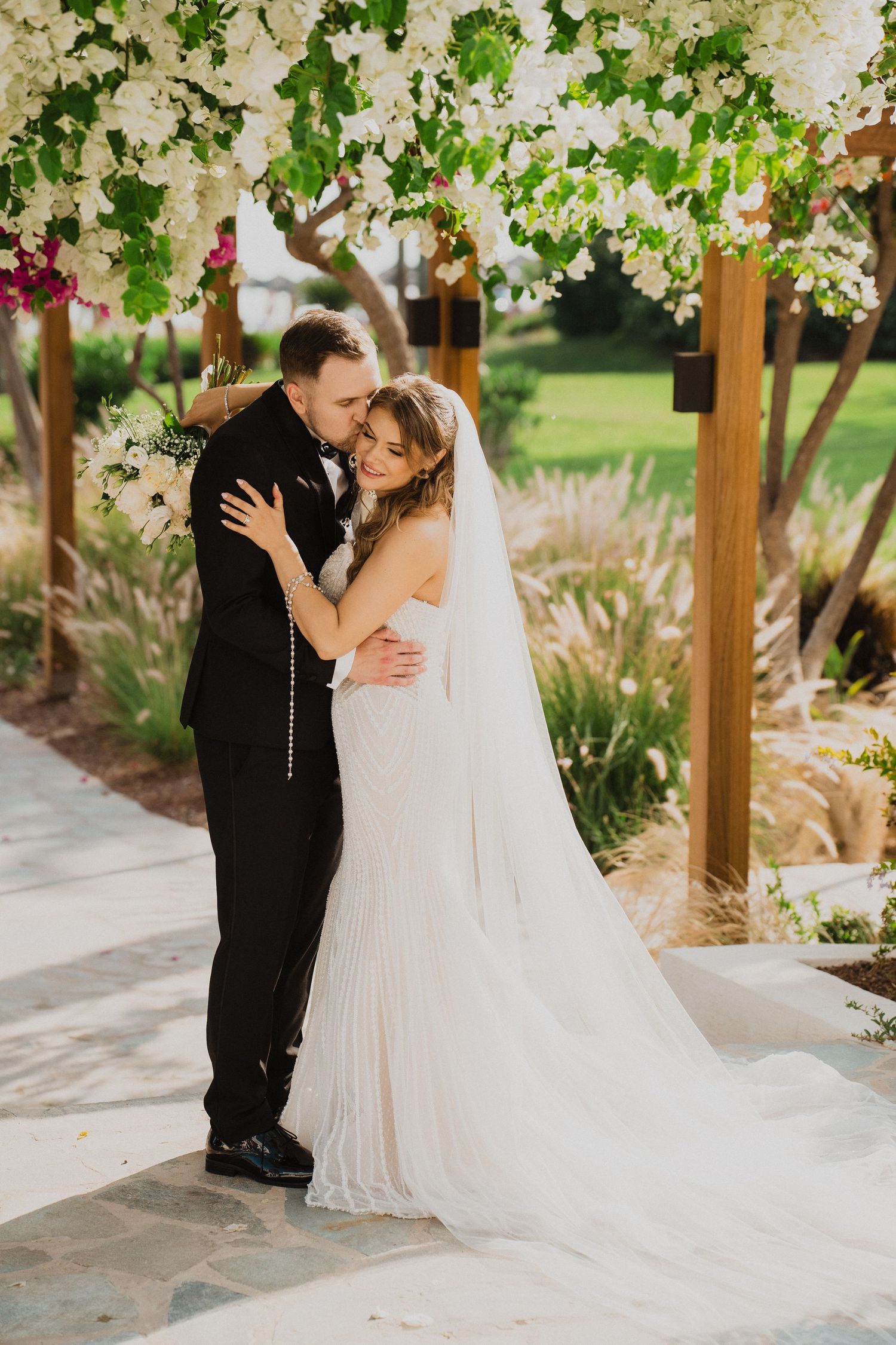 Bride and groom share a romantic embrace under a white floral arch with cascading blooms at outdoor ceremony.
