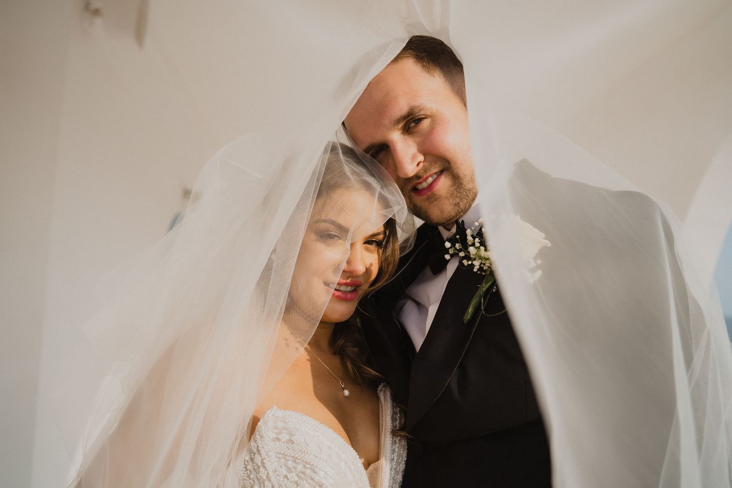 Romantic portrait of bride and groom peeking through a flowing white wedding veil together.