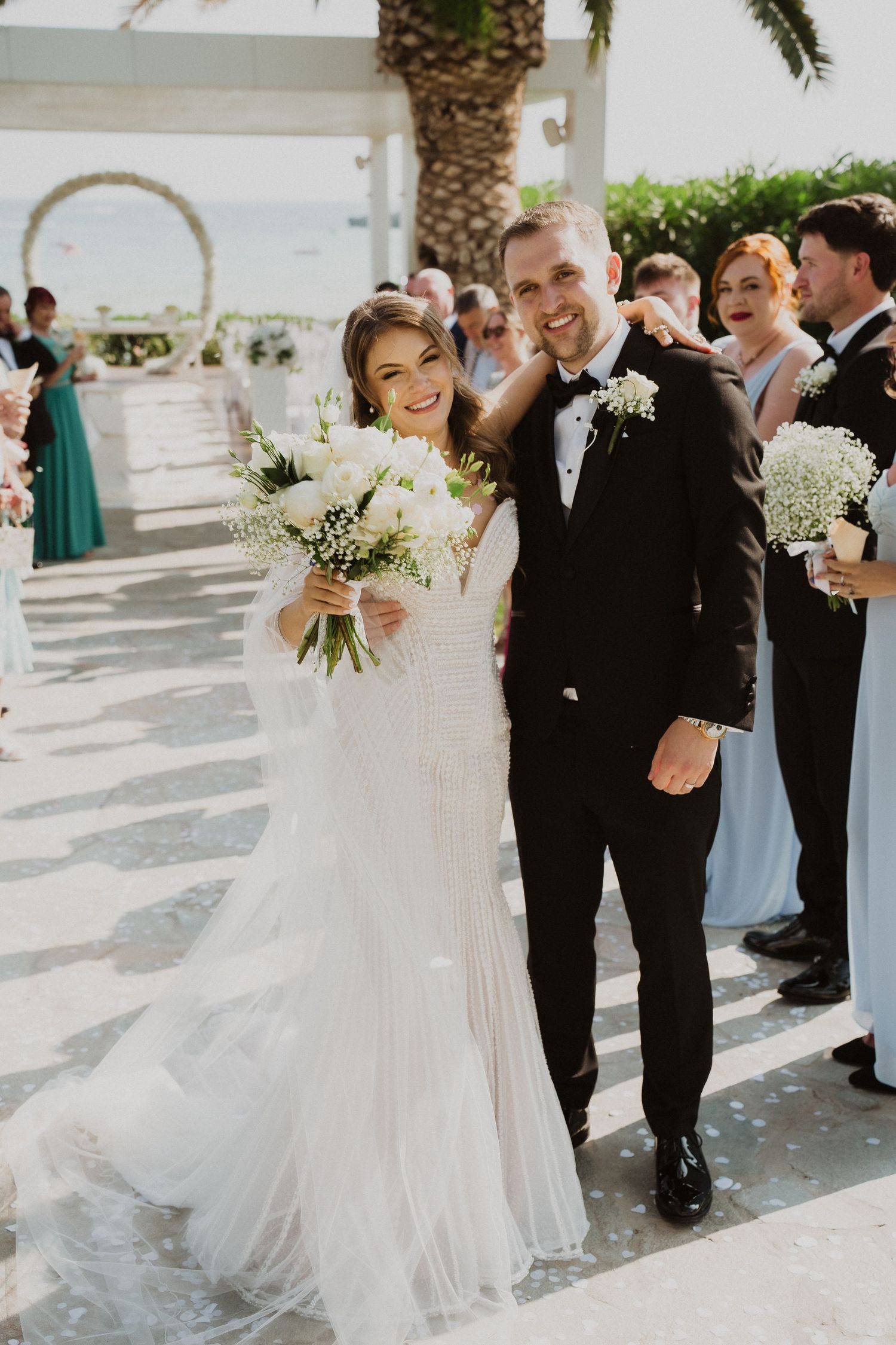 Smiling newlywed couple walks down aisle after wedding ceremony surrounded by cheering guests and palm trees.